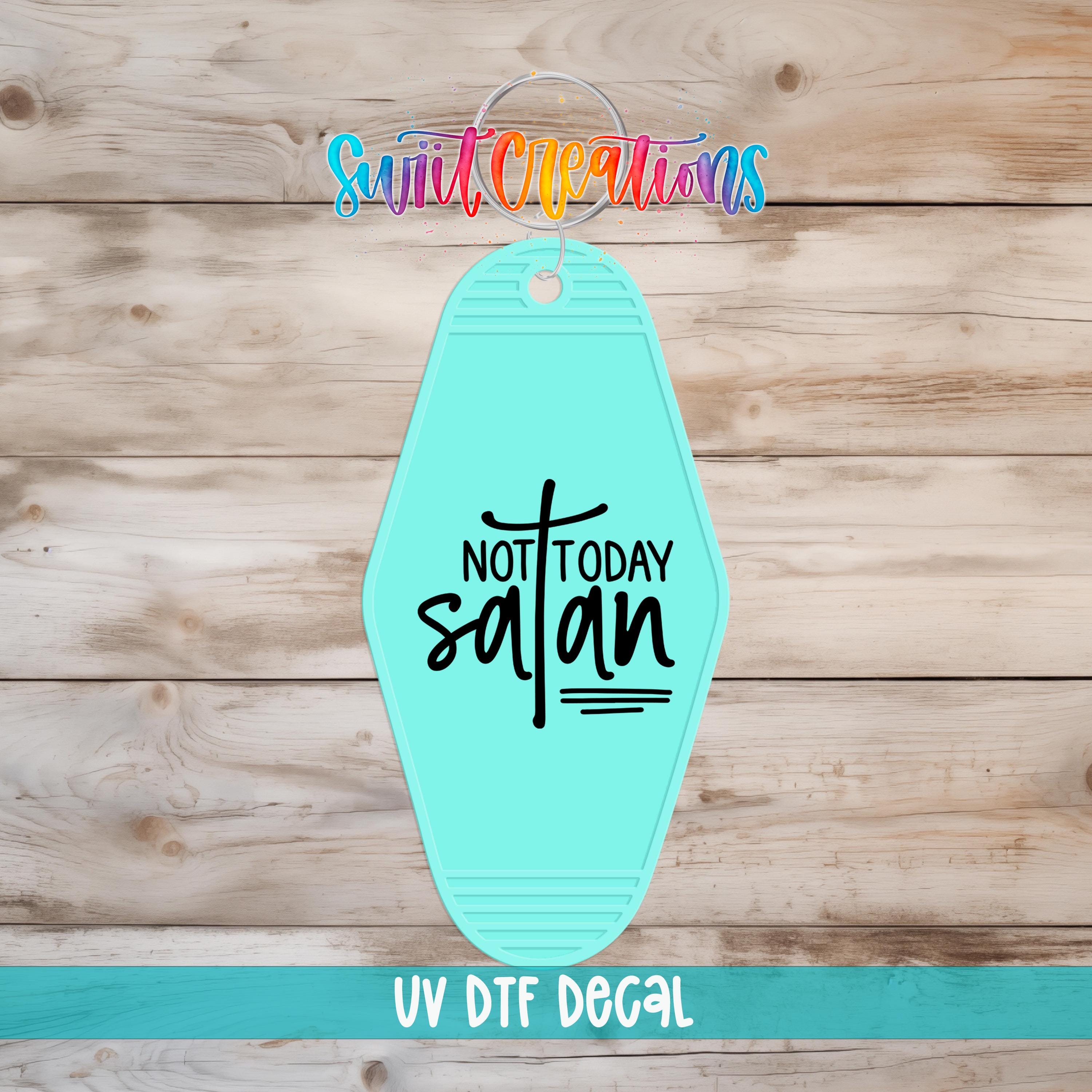 A turquoise-colored keychain with the text "NOT TODAY SATAN" printed on it, placed on a wooden surface.