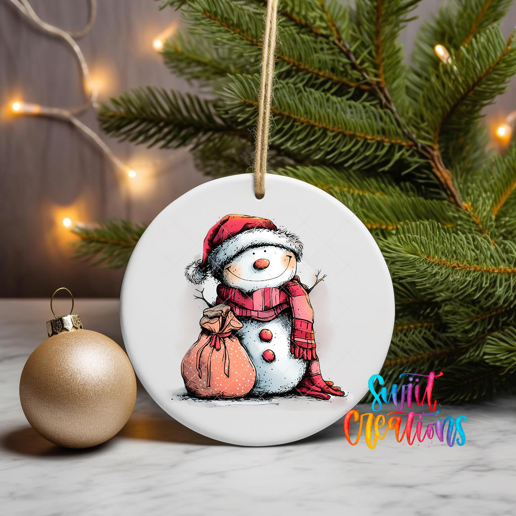 A white ceramic ornament featuring a snowman wearing a red scarf and hat, with a bag around its neck, is displayed on a white surface next to a gold ornament and a Christmas tree.