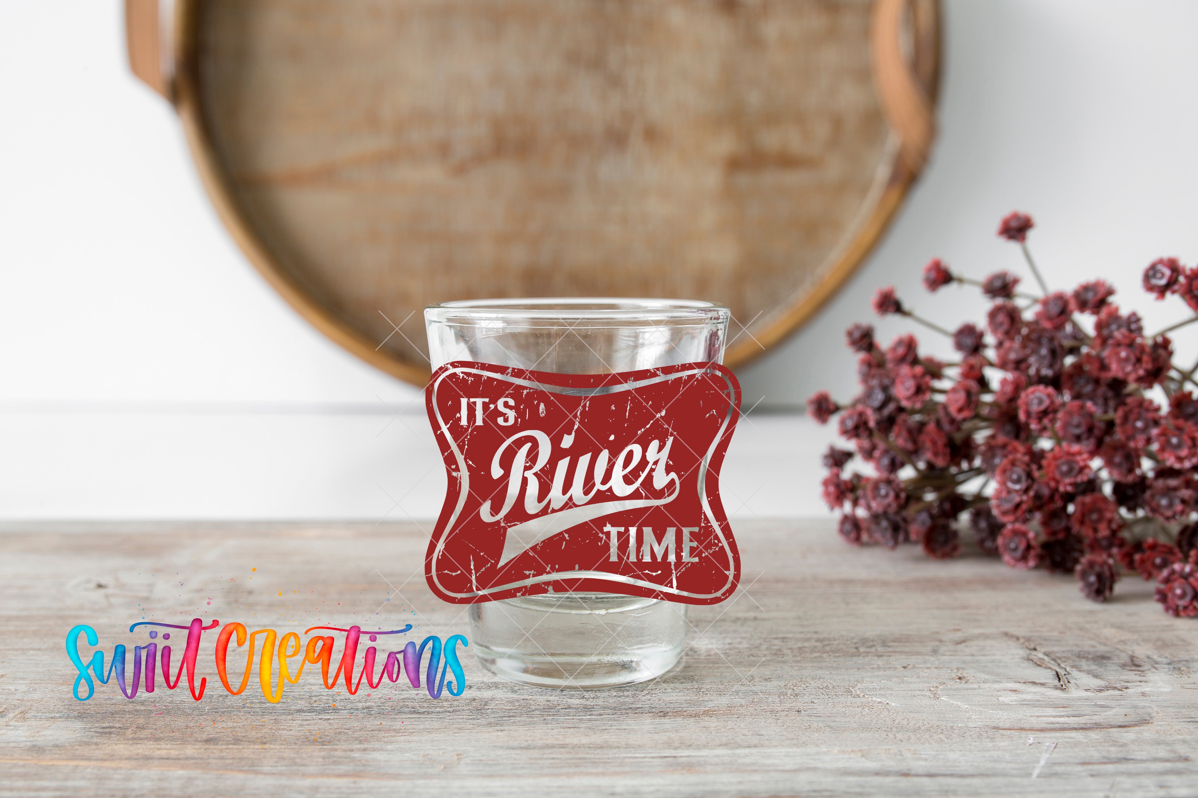 a glass with a red label that says "It's River Time" on a wooden surface, with a bunch of red flowers in the background.