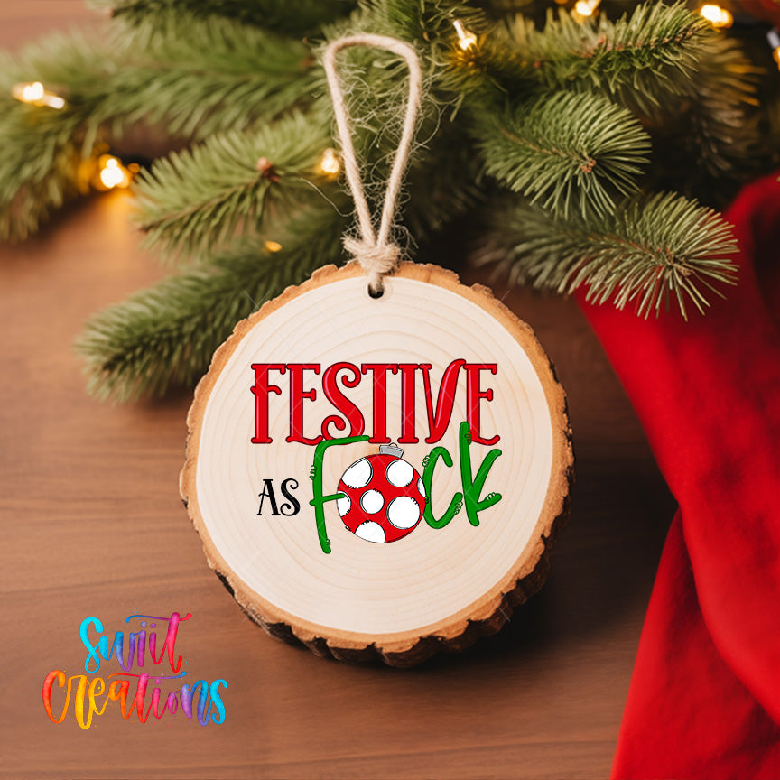 A wooden ornament with the text "FESTIVE AS FOCK" hanging on a Christmas tree, surrounded by pine branches and twinkling lights.
