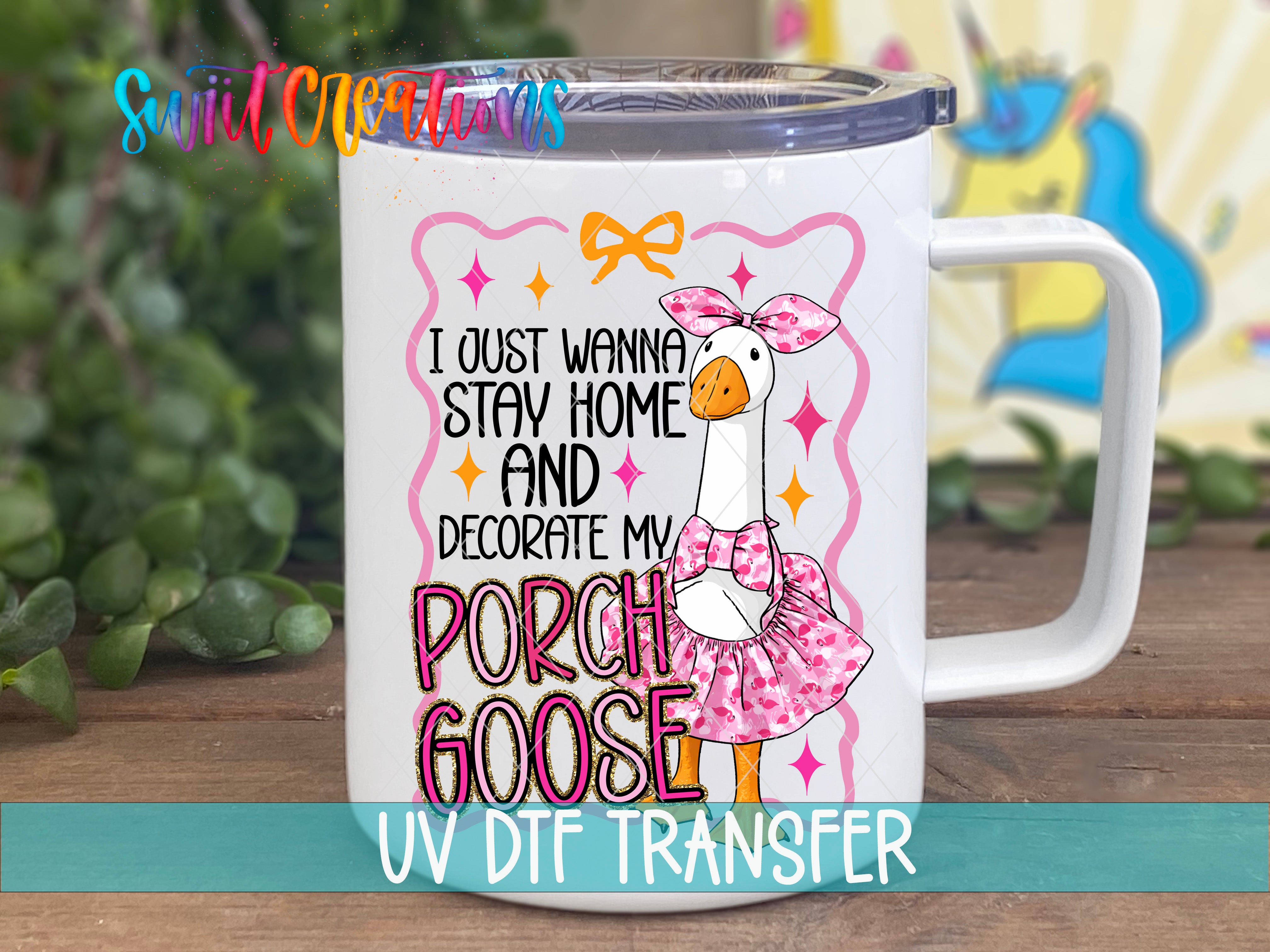 A white mug with a colorful design featuring a cartoon goose wearing a pink dress and bow, along with text that says "I just wanna stay home and decorate my porch goose".