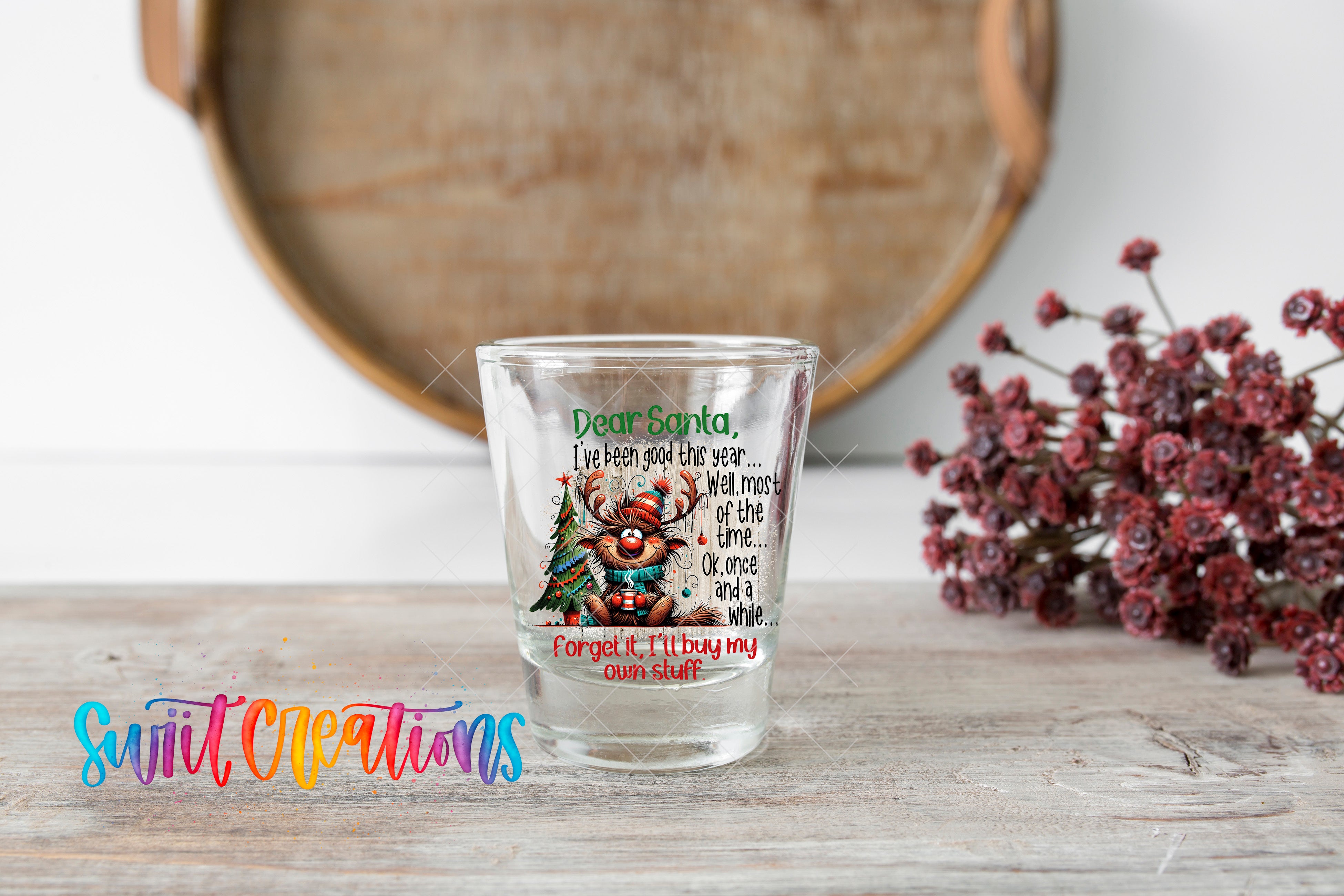 A shot glass with a Santa Claus design and text on it, placed on a wooden surface next to a bunch of red flowers.