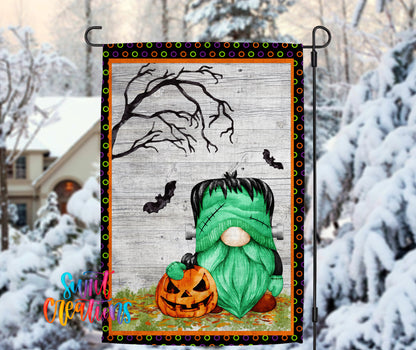 A Halloween-themed garden decoration featuring a green gnome holding a carved pumpkin, with a spooky tree and bats in the background.