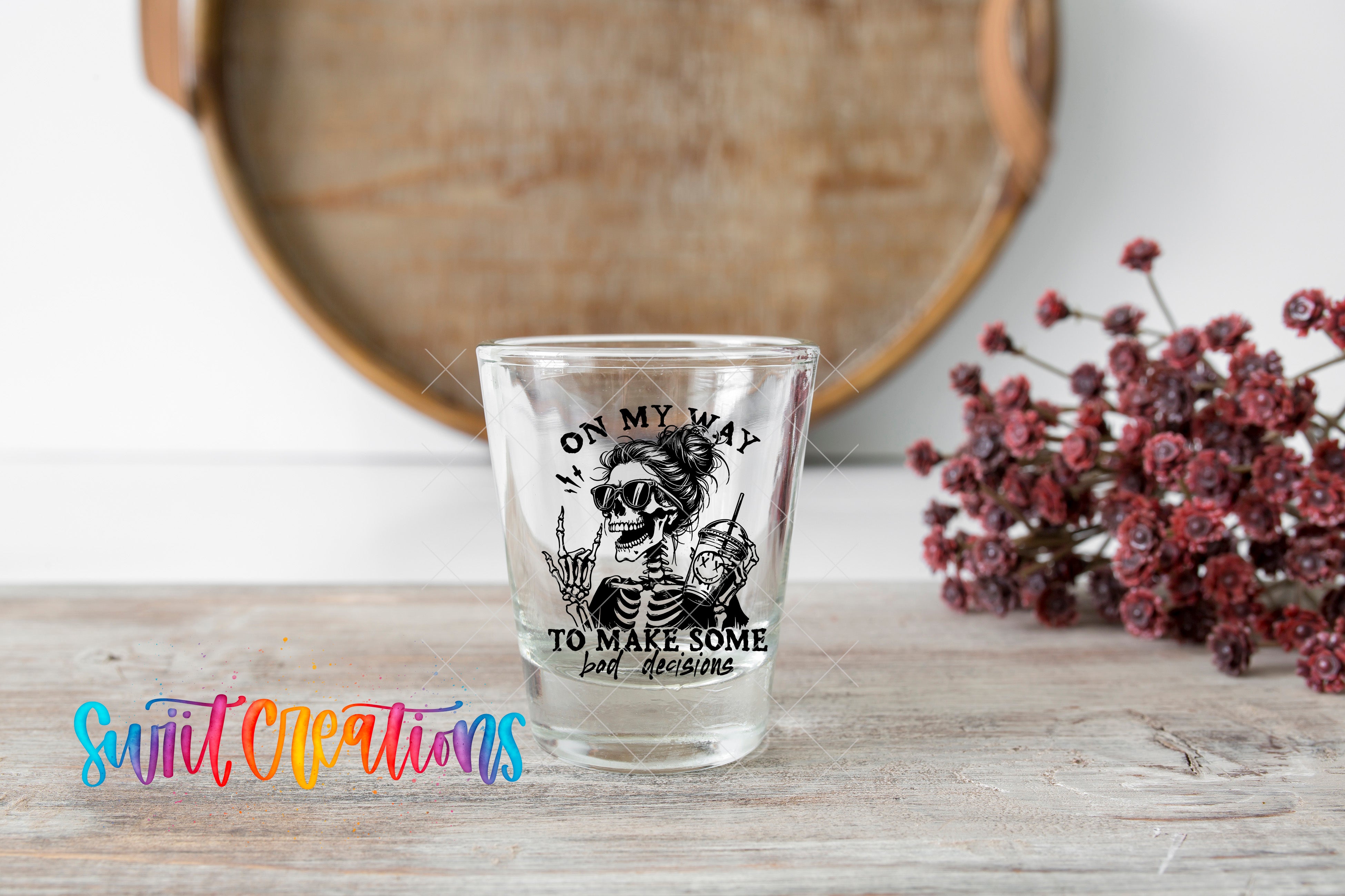 A shot glass with a skeleton design and the text "ON MY WAY TO MAKE SOME BAD DECISIONS" printed on it, placed on a wooden surface next to a bunch of red flowers.