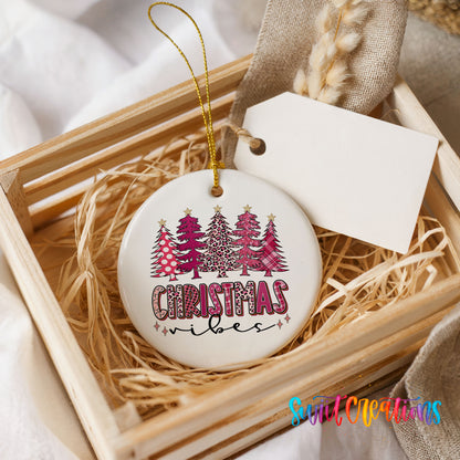 A white ceramic Christmas ornament with a pink and black tree design, placed in a wooden crate filled with straw.