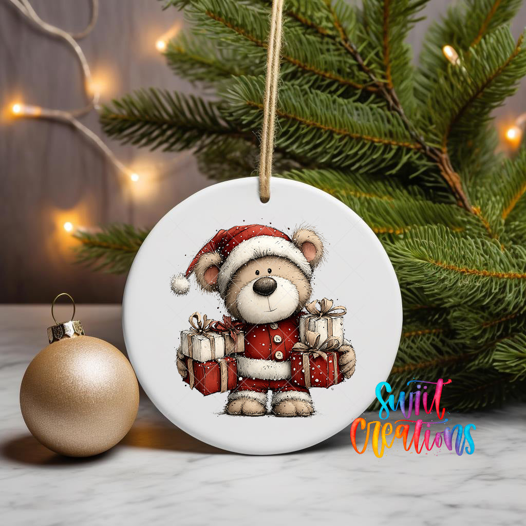 A white ceramic ornament featuring a teddy bear wearing a Santa hat and holding presents, placed on a marble surface next to a gold ornament and a Christmas tree.