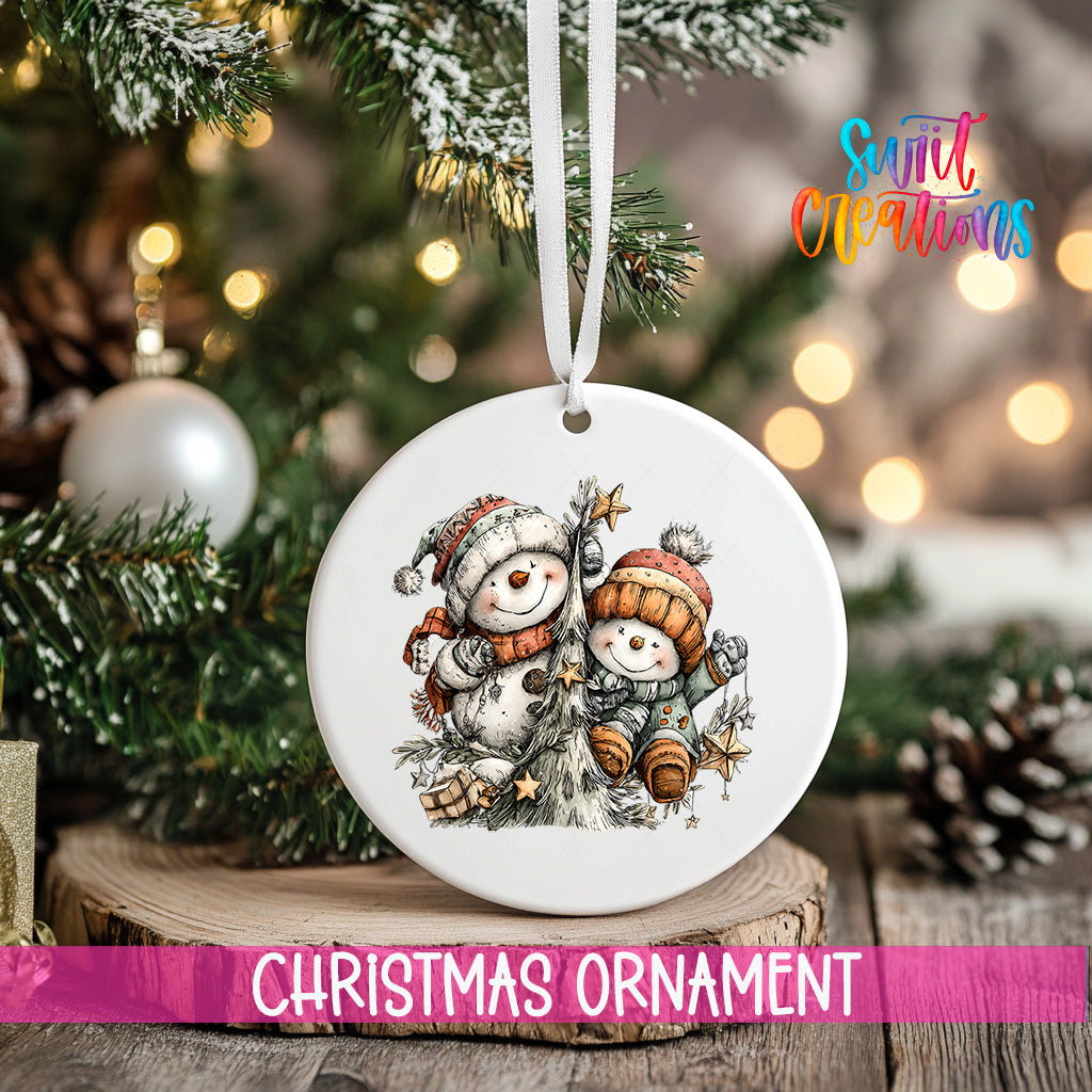 A white Christmas ornament featuring two snowmen is hanging from a tree branch, with a blurred background of a decorated Christmas tree and other ornaments.