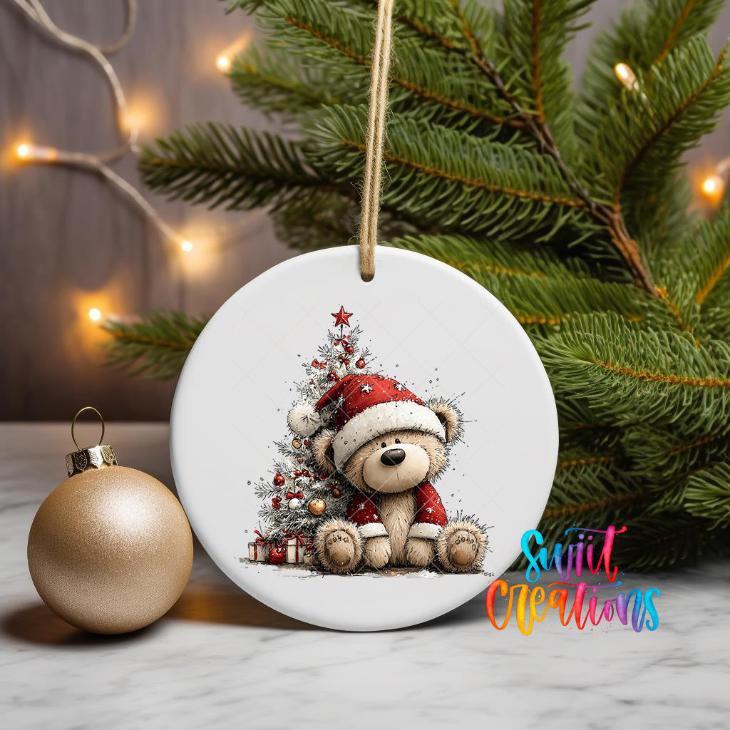 A white ceramic ornament featuring a teddy bear wearing a Santa hat, sitting in front of a Christmas tree, is displayed on a table with a gold ornament and a string of lights in the background.
