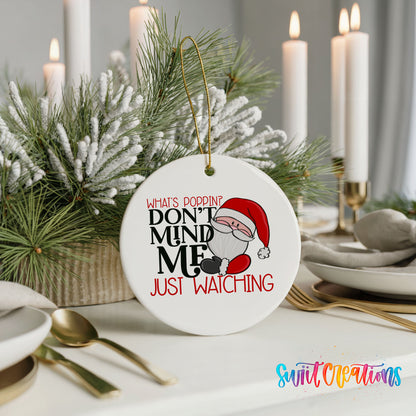A white ceramic ornament with a Santa Claus design and text that reads "What's POPPIN? DON'T MINDS, DON'T MIND ME, JUST WATCHING". The ornament is placed on a table with a candle and other holiday decorations.