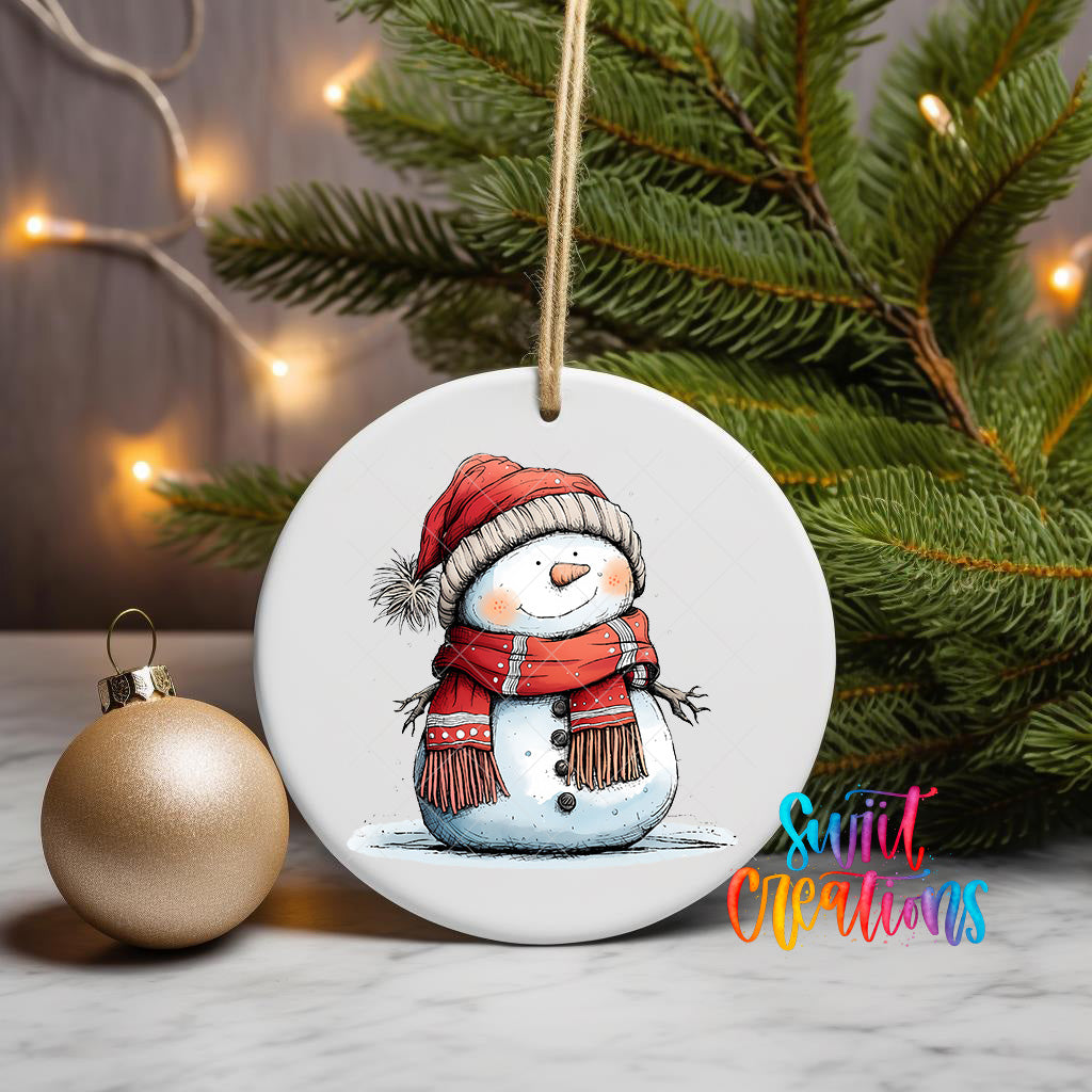 A white ceramic ornament featuring a snowman wearing a red scarf and hat, with a Christmas tree and twinkling lights in the background.