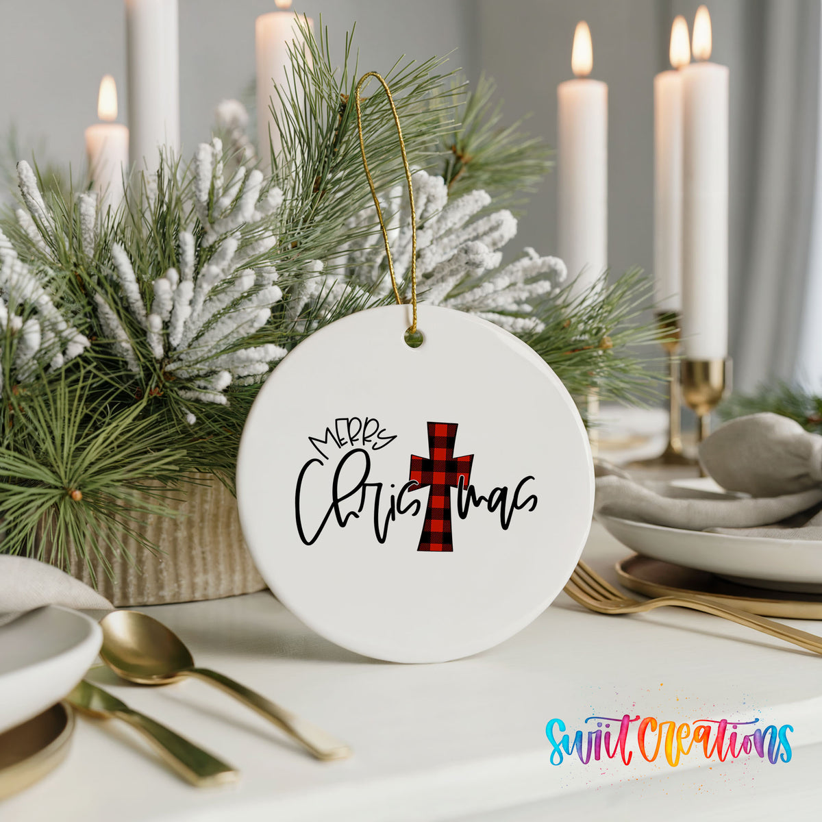 A white Christmas ornament with a red and black plaid cross design hangs on a table, surrounded by a festive arrangement of pine branches and candles.