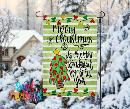 A festive Christmas-themed flag is displayed in a snowy outdoor setting, featuring a tree with red and green ornaments and a star on top.