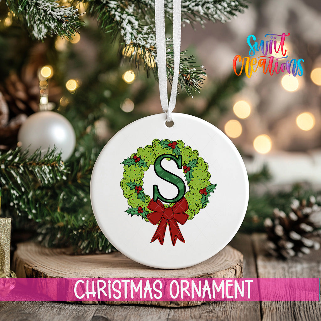 A white Christmas ornament with a green wreath and a red ribbon is hanging from a tree branch, with a blurred background of lights and pine cones.