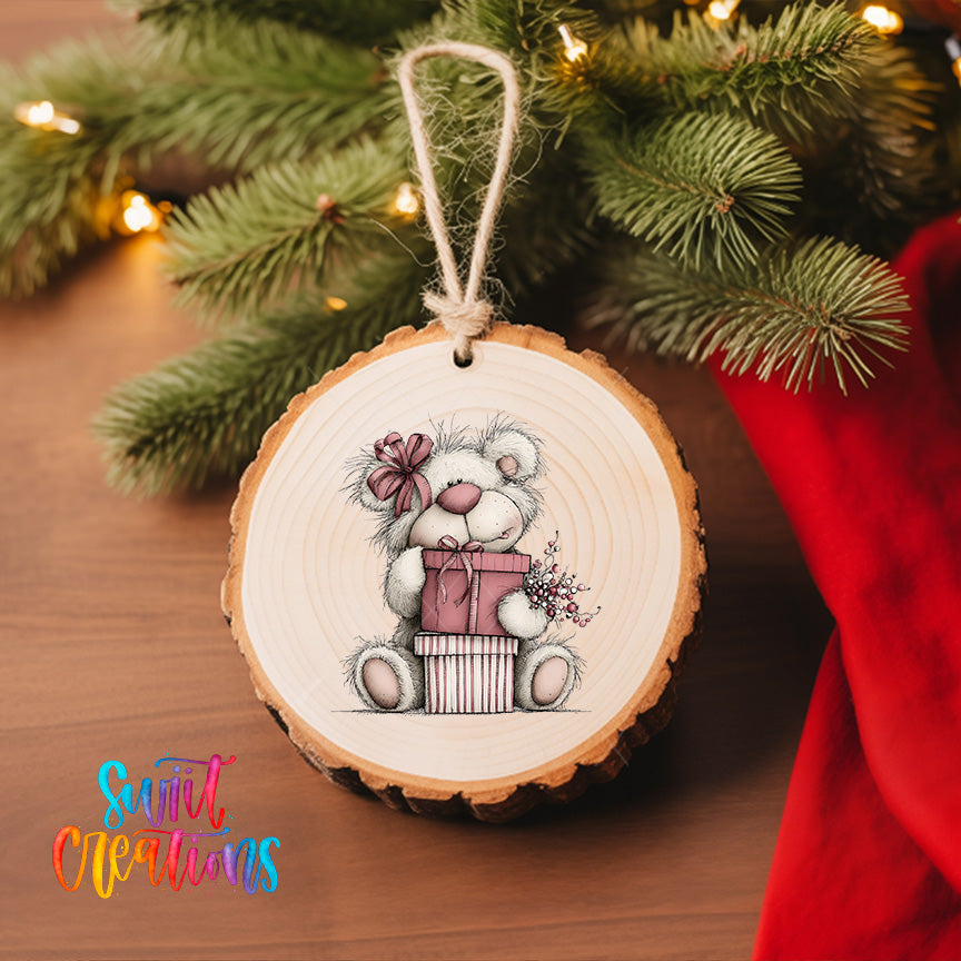 A wooden ornament featuring a teddy bear holding a gift, hanging on a Christmas tree with a red cloth visible in the background.