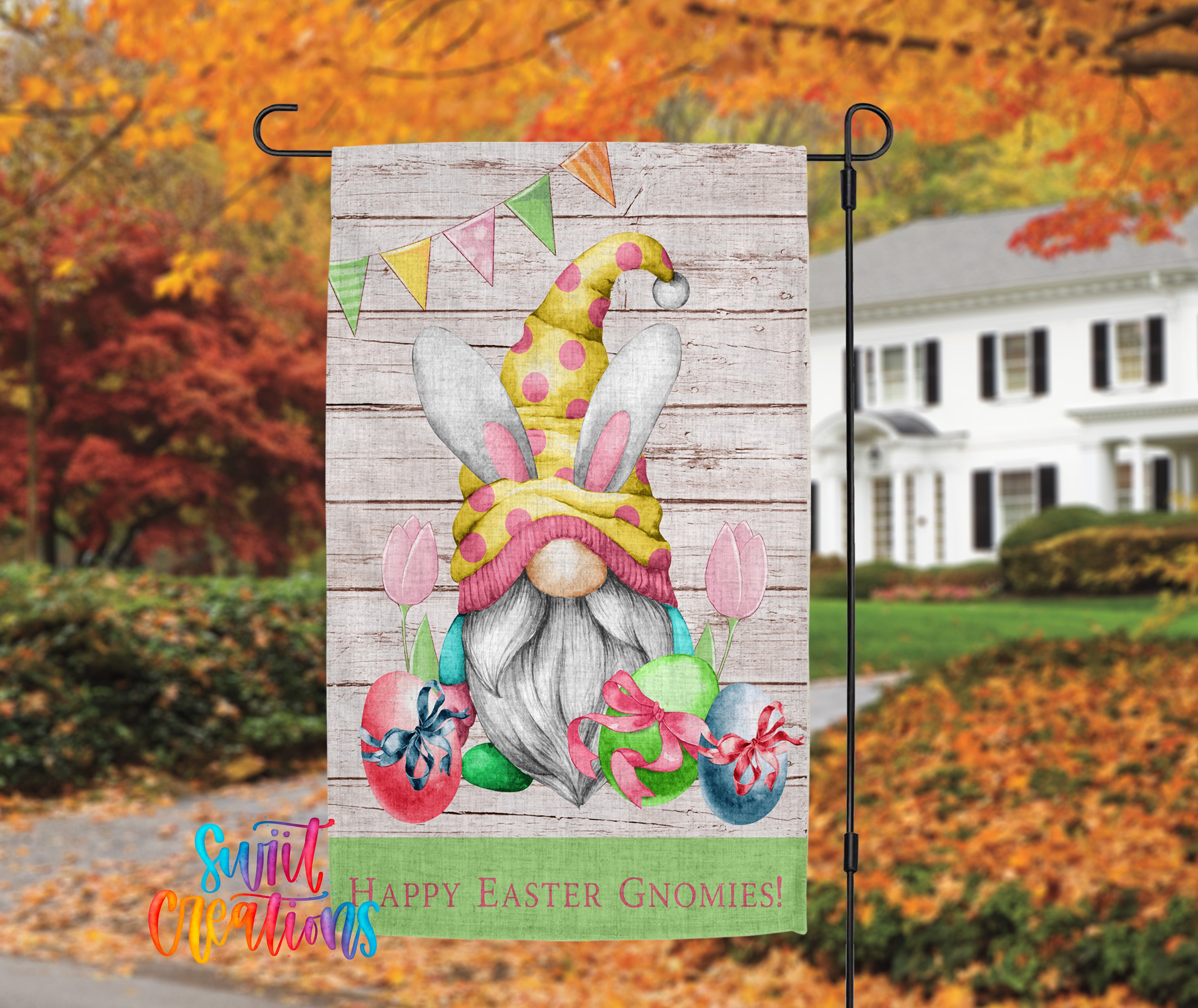 A colorful Easter-themed garden decoration featuring a gnome wearing a bunny hat, surrounded by flowers and eggs, with a house and autumn trees in the background.