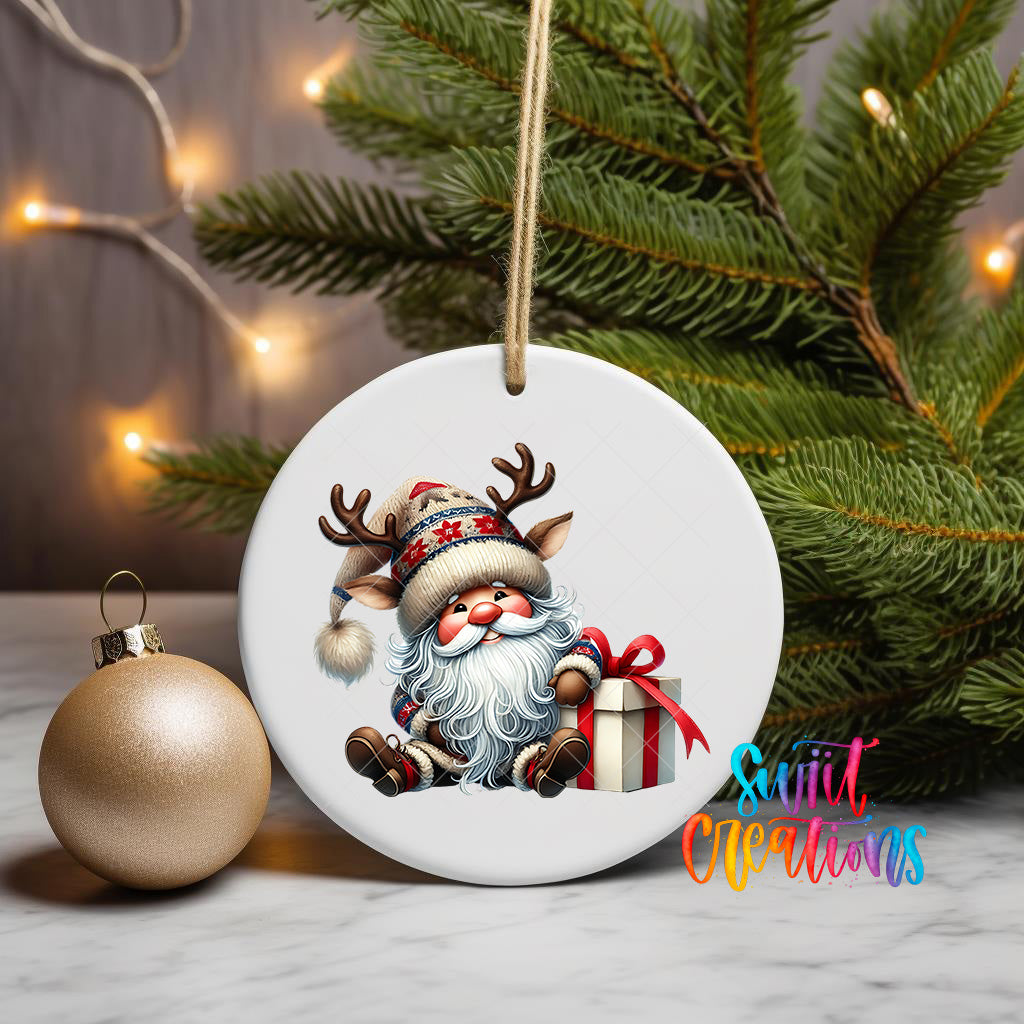 A white ornament featuring a cartoon Santa Claus holding a gift and wearing a reindeer hat, placed on a table next to a gold ornament and a Christmas tree.