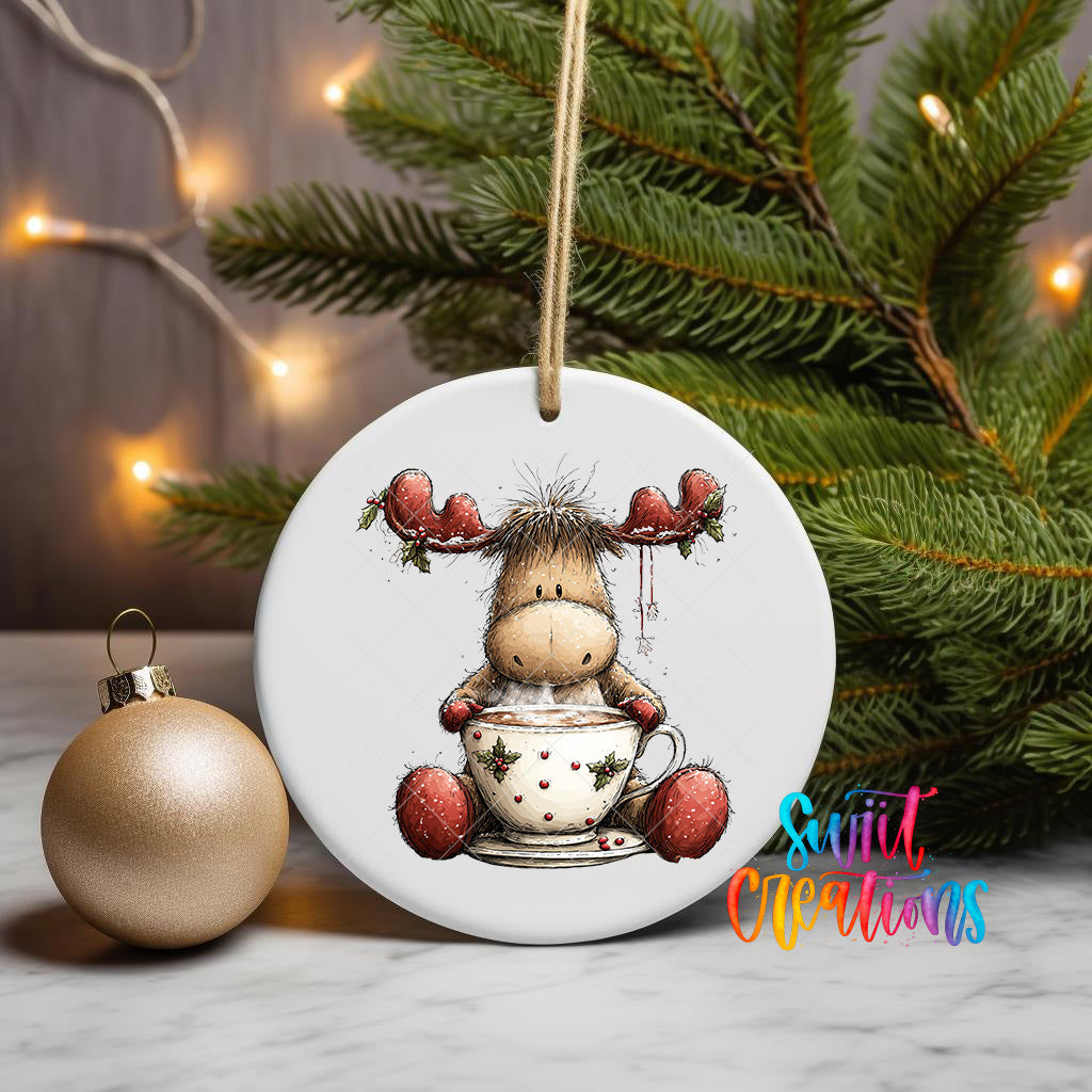 A white ceramic ornament featuring a cartoon moose holding a cup of hot chocolate, surrounded by Christmas lights and greenery.
