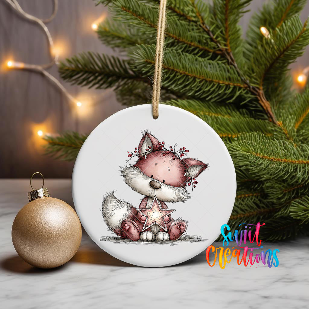 A white ceramic ornament featuring a cartoon image of a fox holding a star, placed on a marble surface next to a gold ornament and a Christmas tree with twinkling lights.
