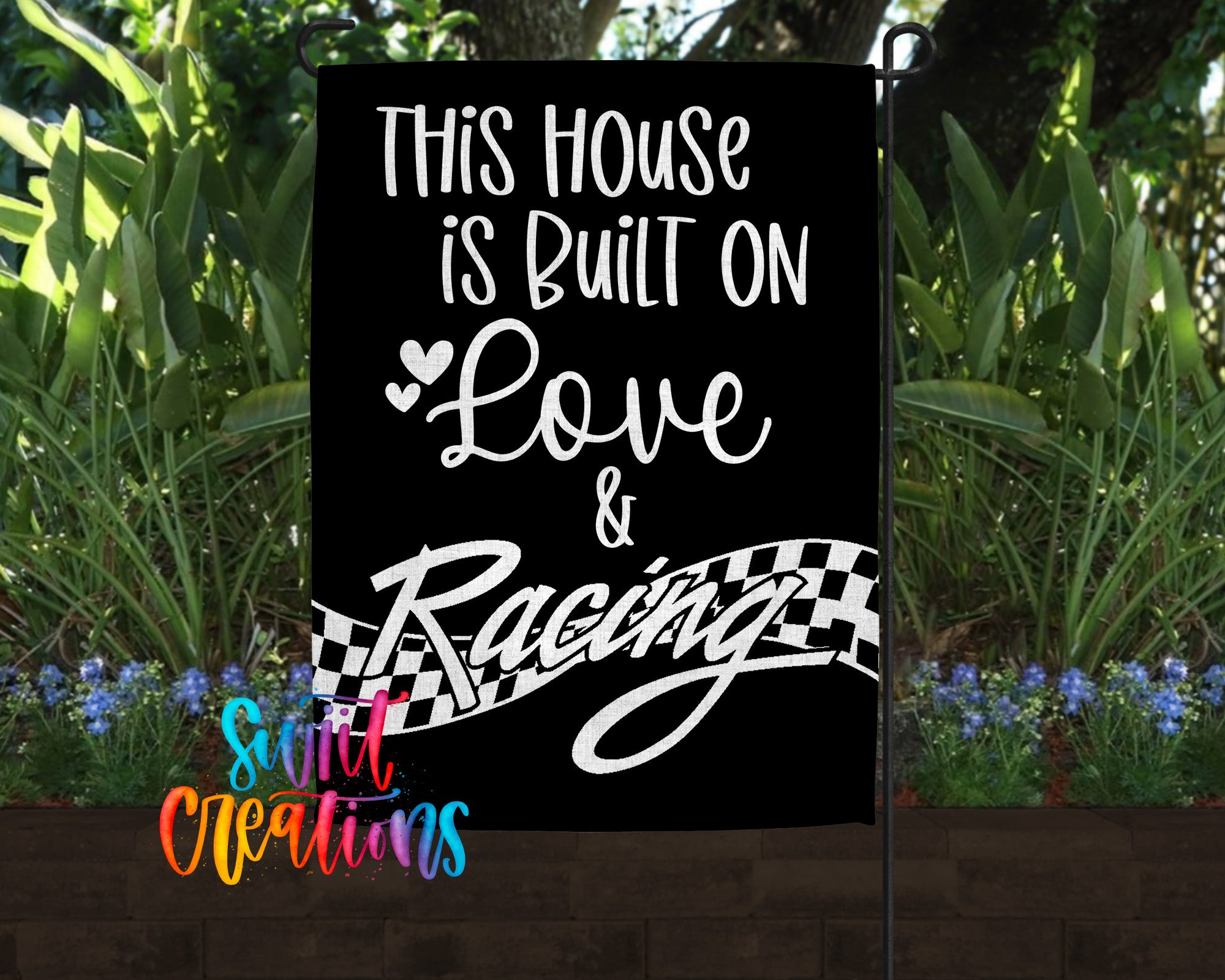 a black and white sign with the text "This house is built on love & racing" displayed on it, set against a backdrop of lush greenery and flowers.