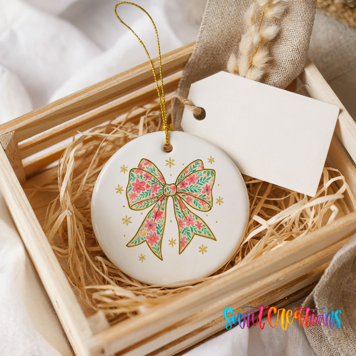 A white ceramic ornament with a floral design and a gold bow is displayed in a wooden crate, accompanied by a white tag.