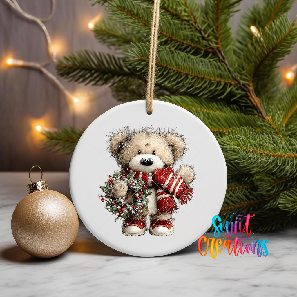 A white ceramic ornament featuring a teddy bear wearing a red scarf and holding a wreath, placed on a marble surface next to a gold ornament and a Christmas tree with twinkling lights.