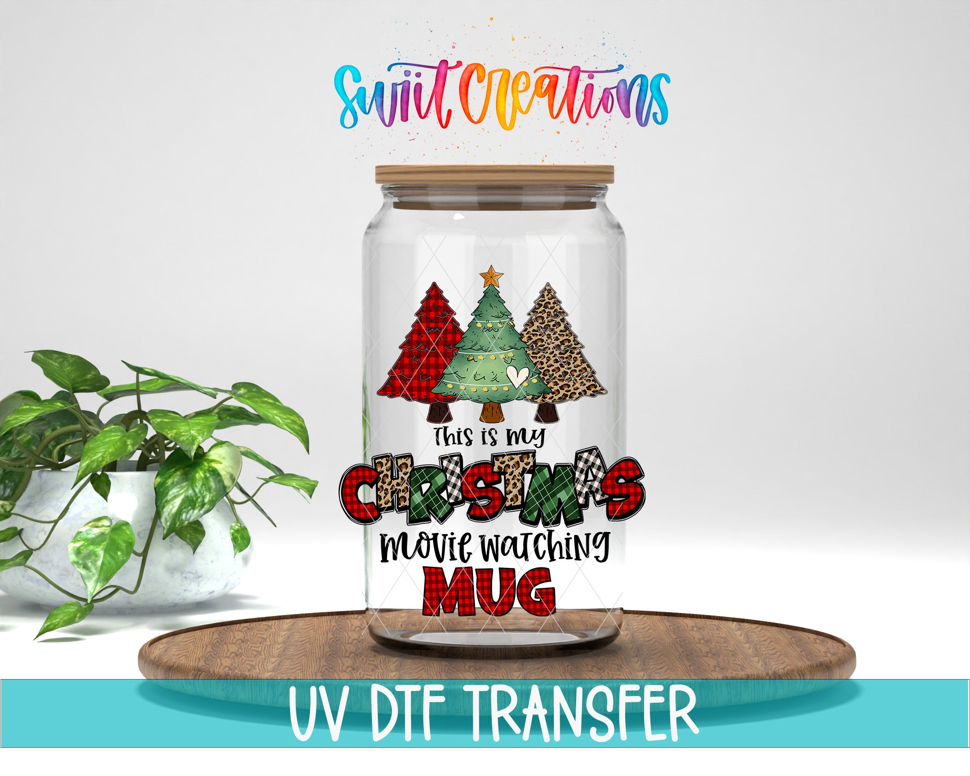 A glass jar with a Christmas tree design and text, placed on a wooden surface next to a potted plant.