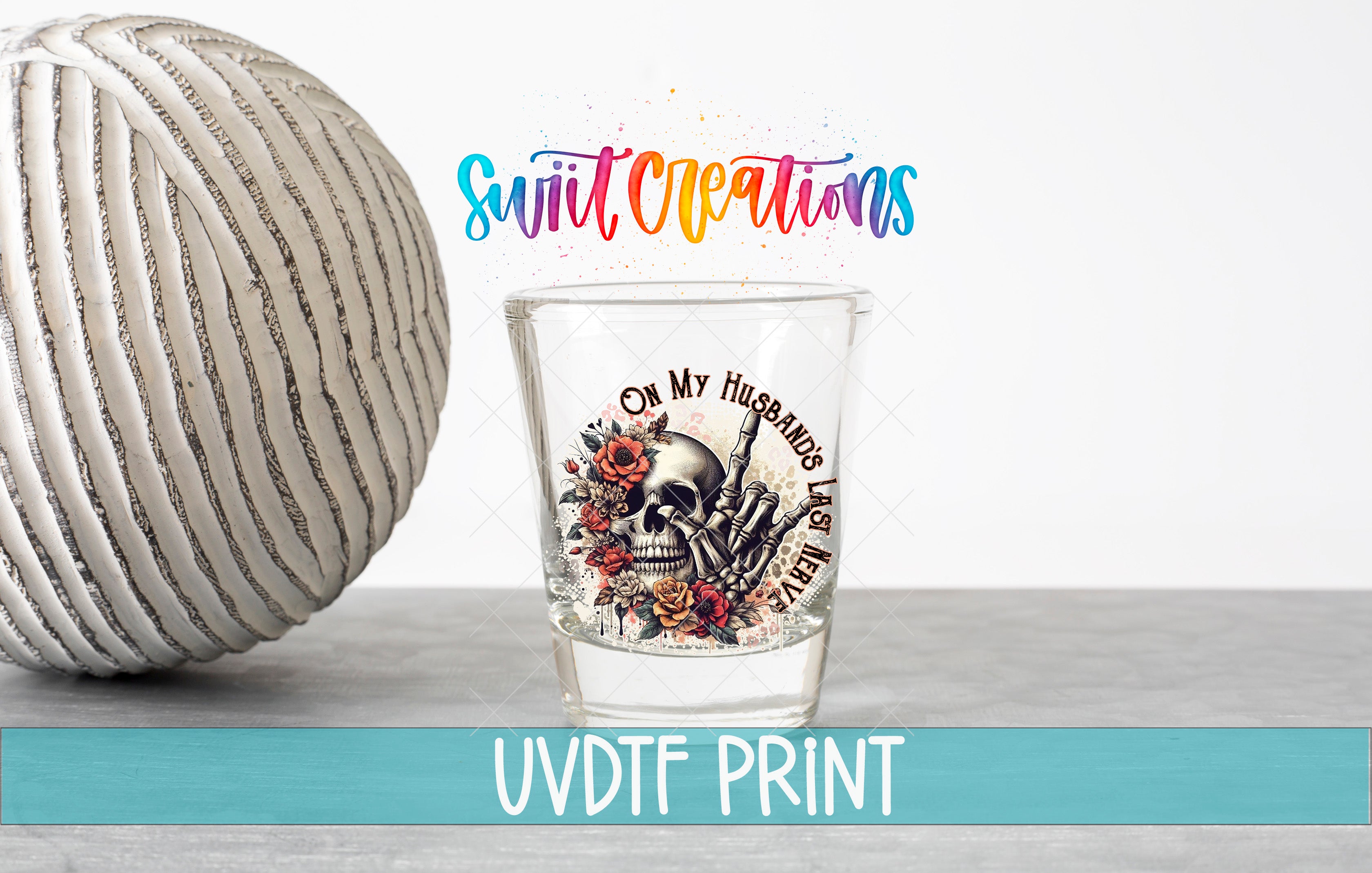 a shot glass with a skull design and floral elements, placed on a table next to a white ball.