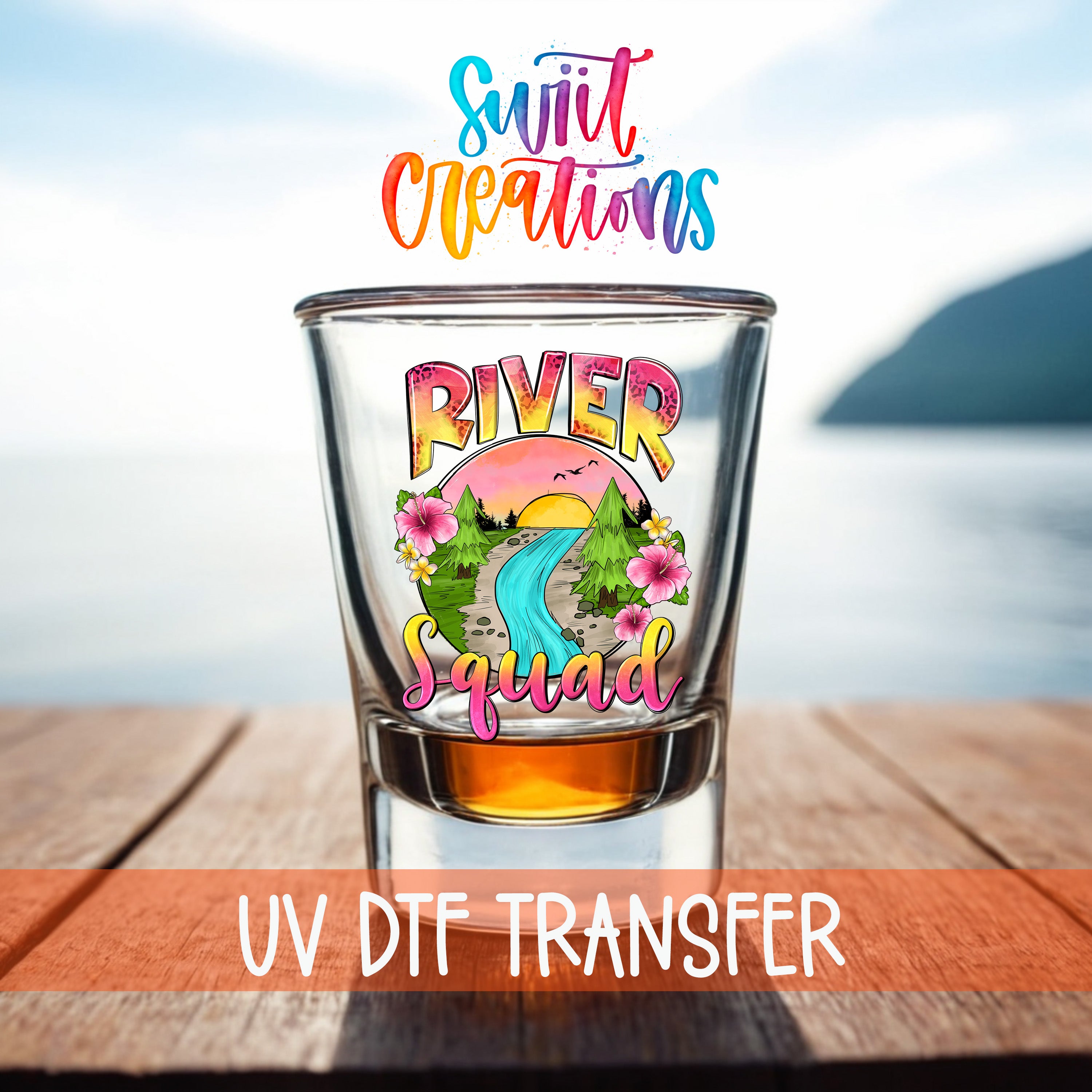 A shot glass with a colorful design featuring a river and flowers, placed on a wooden surface with a body of water in the background.