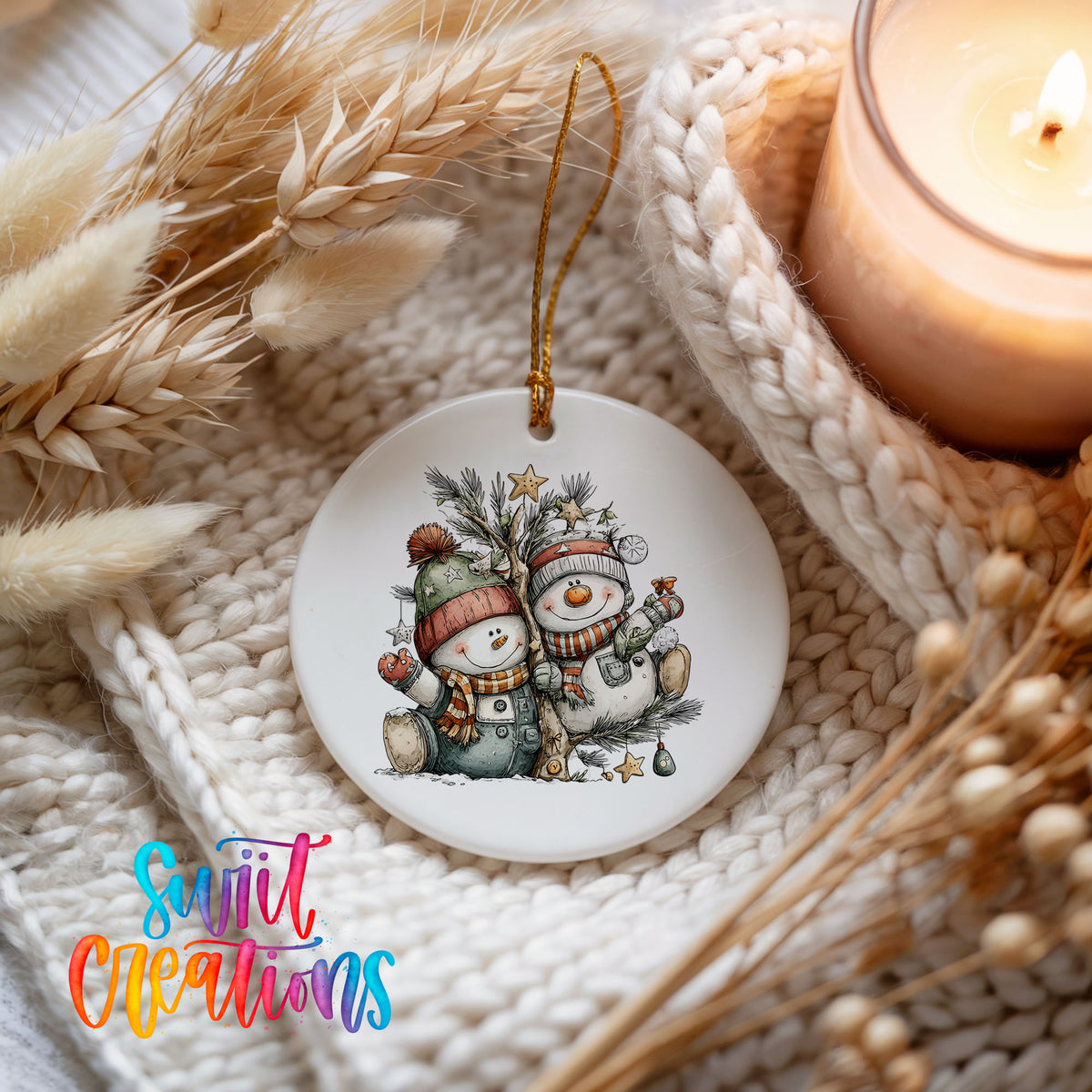 A white ceramic ornament featuring two snowmen, one wearing a green hat and the other a red hat, sitting together on a branch. The ornament is hanging from a string, and there is a lit candle and some dried wheat stalks in the background.