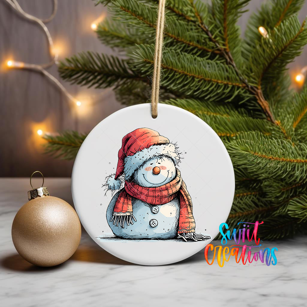 A white ceramic ornament featuring a snowman wearing a red scarf and hat, sitting on a white surface next to a gold ornament and a Christmas tree with twinkling lights.