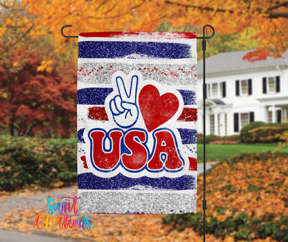 A patriotic flag with the word "USA" prominently displayed, featuring a peace sign and a red heart symbol. The flag is hanging on a black pole, with a house and autumn foliage visible in the background.