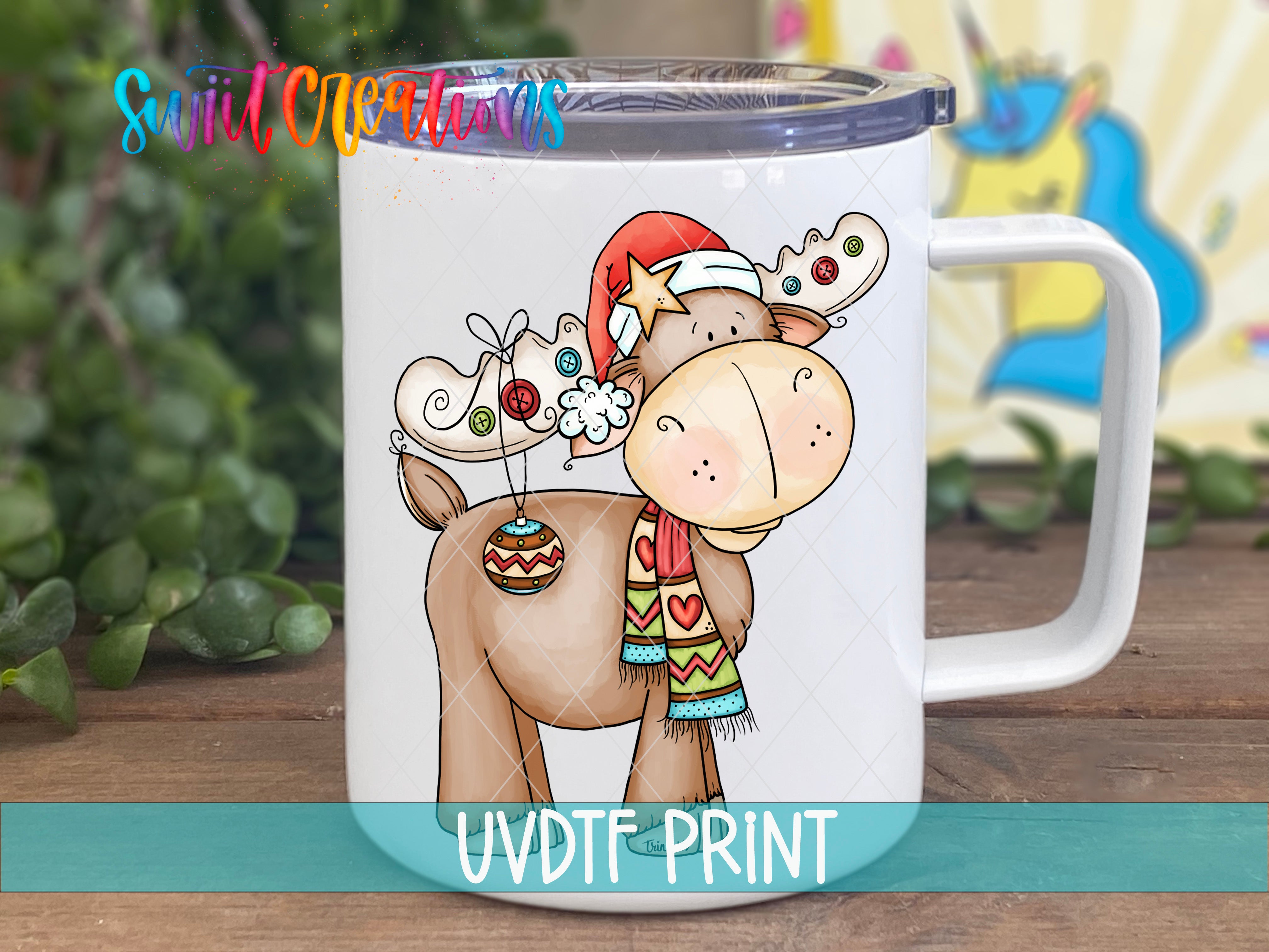 A mug with a cartoon moose wearing a Santa hat and scarf, along with other festive decorations, is displayed on a wooden surface.