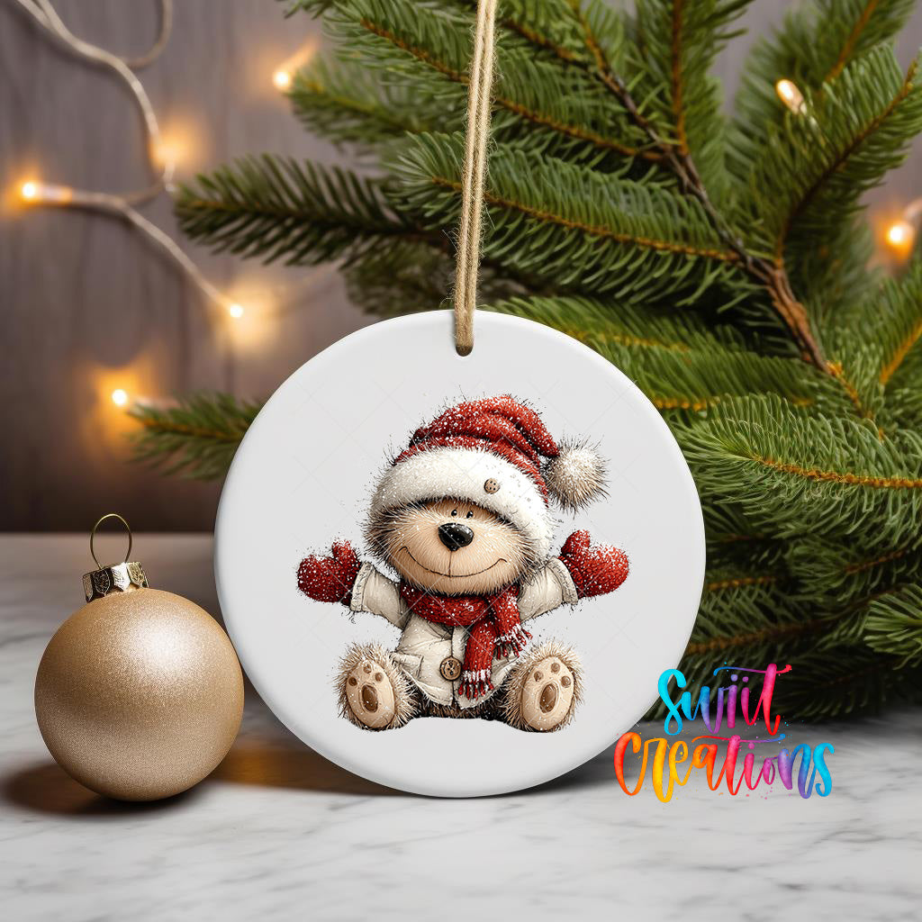 A white ceramic ornament featuring a teddy bear wearing a Santa hat and mittens, sitting on a white surface next to a gold ornament and a Christmas tree with twinkling lights.