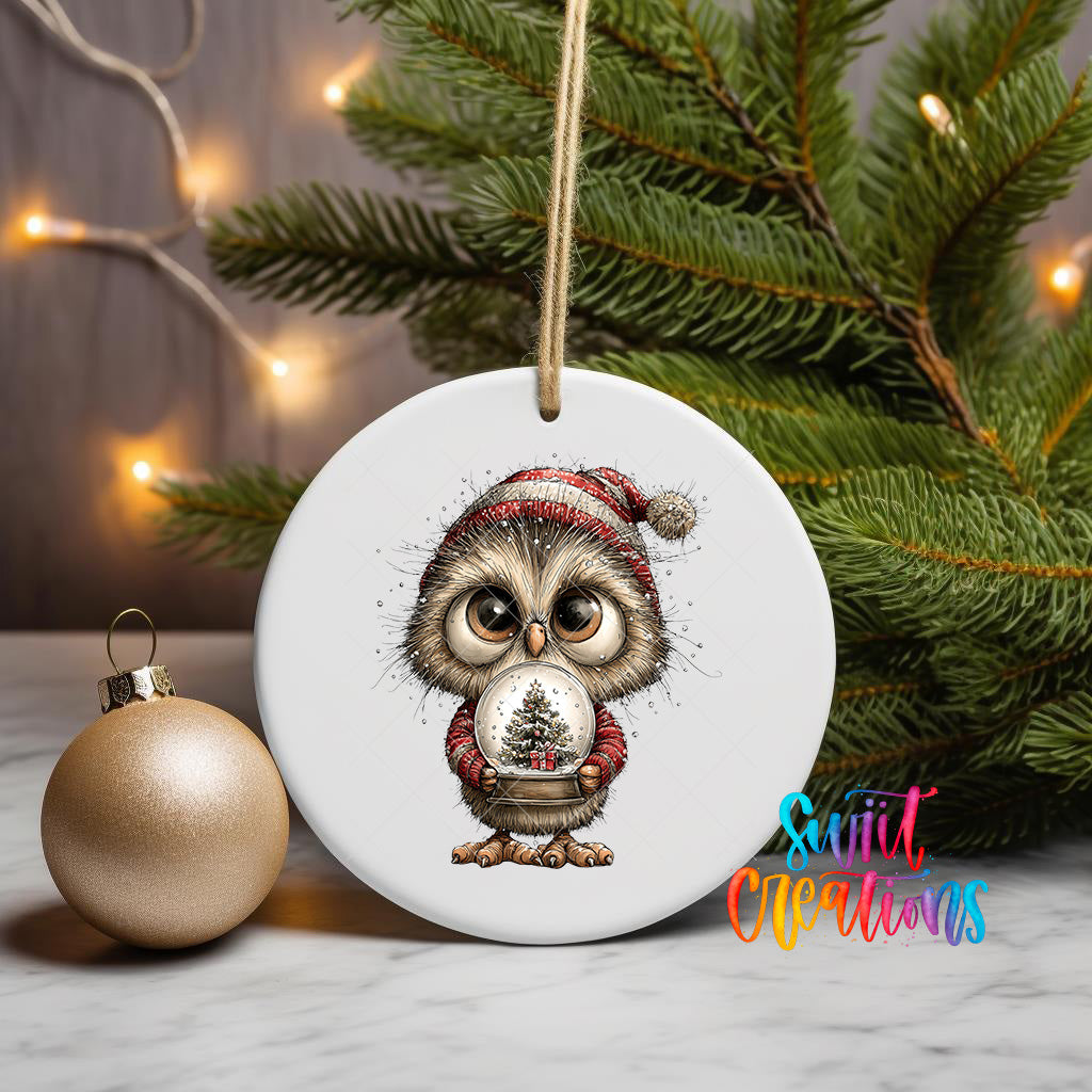 A white ceramic ornament featuring a cartoon owl wearing a red and white striped hat, holding a snow globe, is displayed on a white surface next to a gold ornament and a Christmas tree.