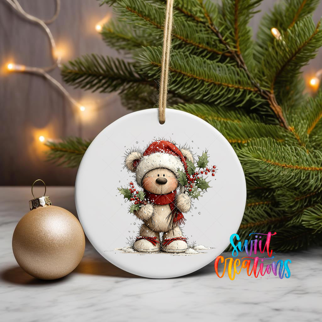 A white ceramic ornament featuring a teddy bear wearing a Santa hat and scarf, holding a holly bush, is displayed on a white marble surface next to a gold ornament and a Christmas tree.