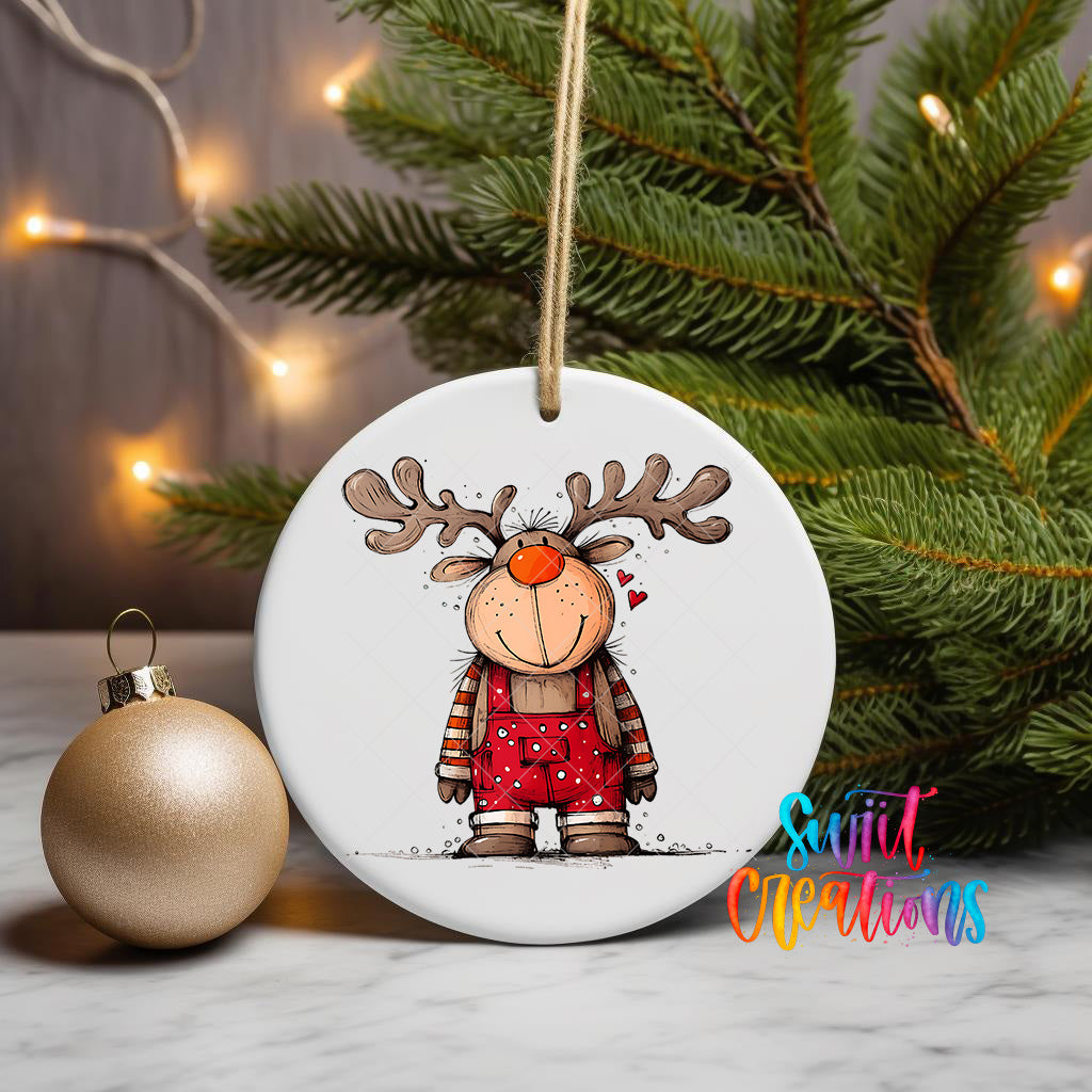 A white ceramic ornament featuring a cartoon reindeer with antlers and a red scarf, hanging on a string next to a gold ornament and a Christmas tree.