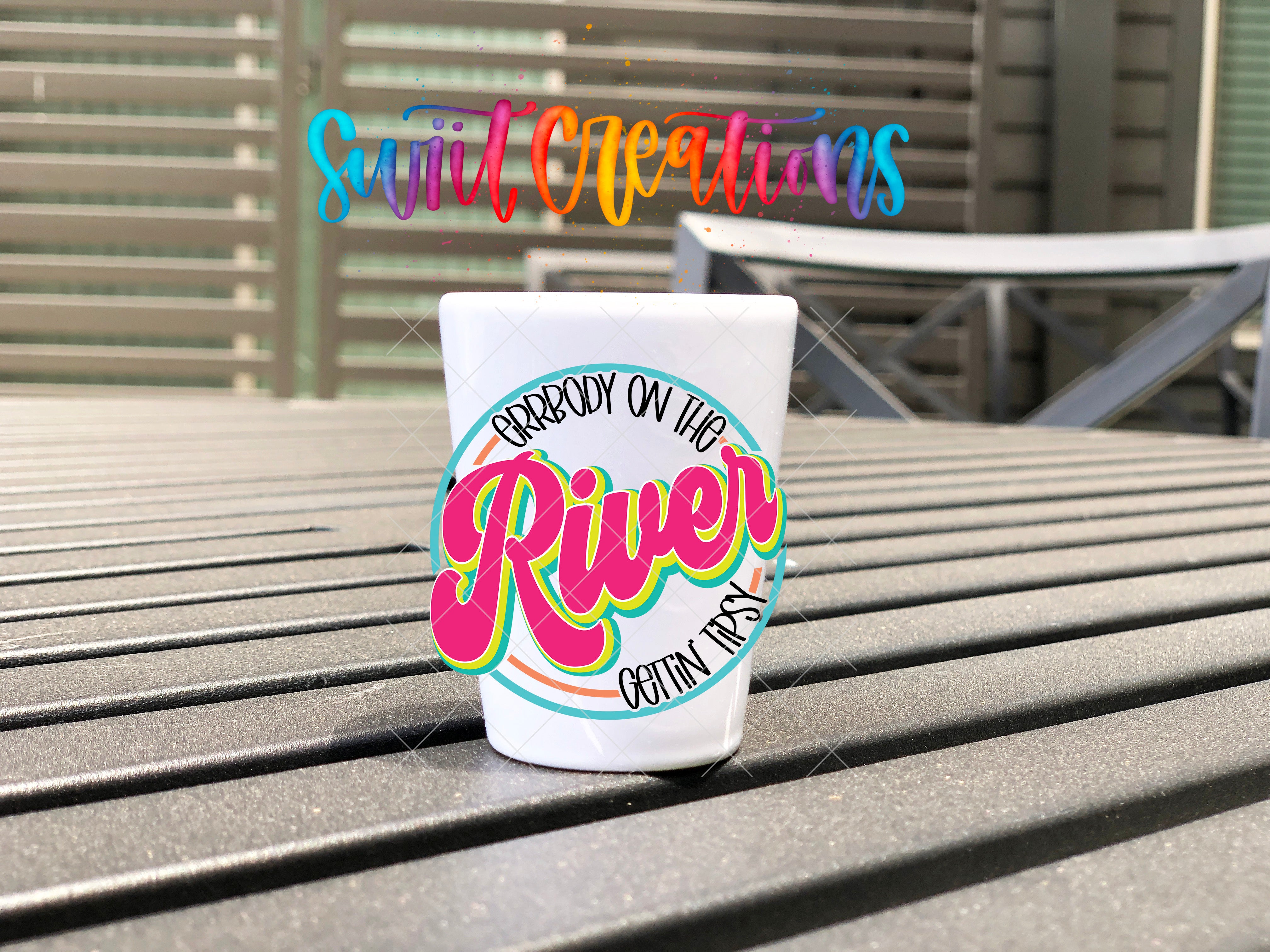 A white cup with a colorful design sits on a wooden surface, with the text "River" and "Arrbody on the River" displayed on the cup.