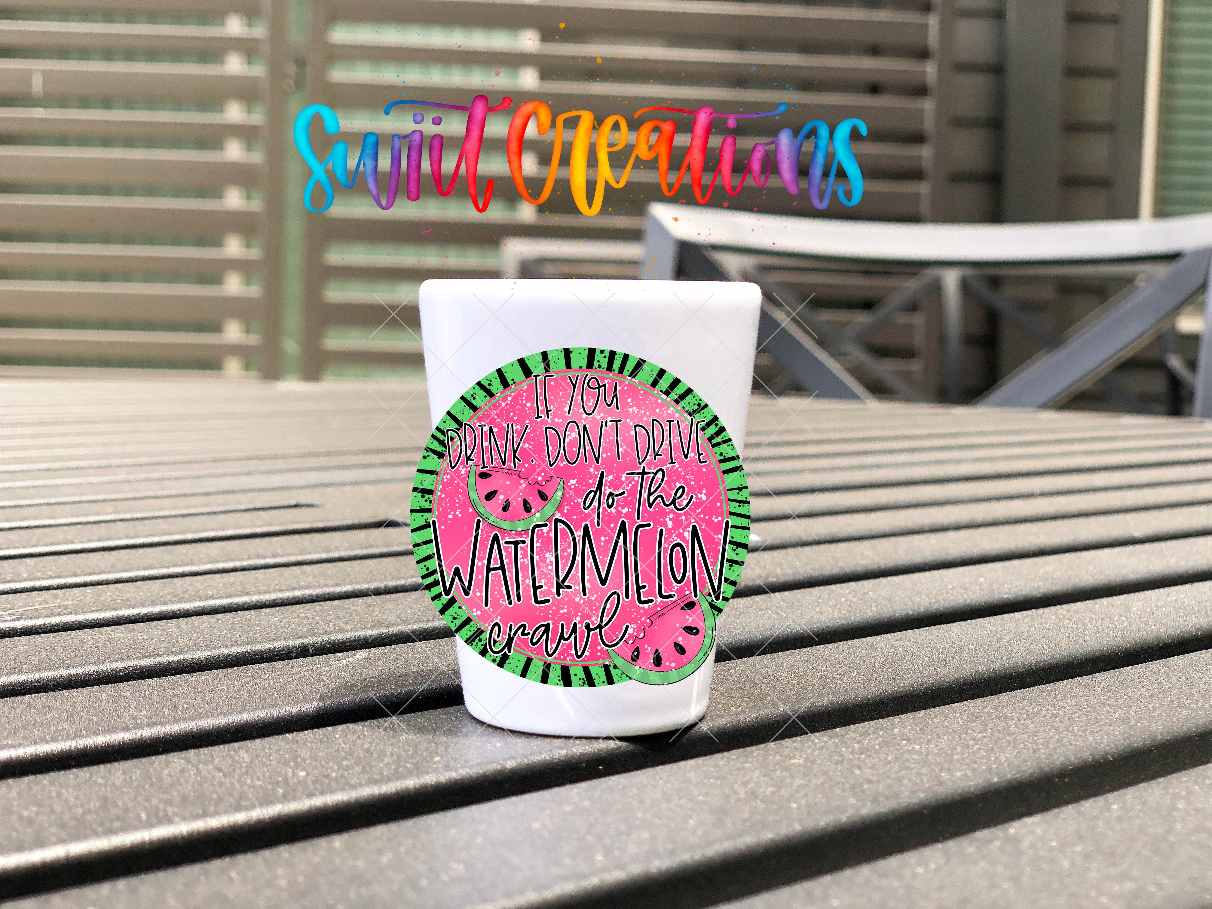 A white cup with a colorful design featuring a watermelon and the text "If you drink don't drive on the watermelon crawl" is placed on a wooden surface.