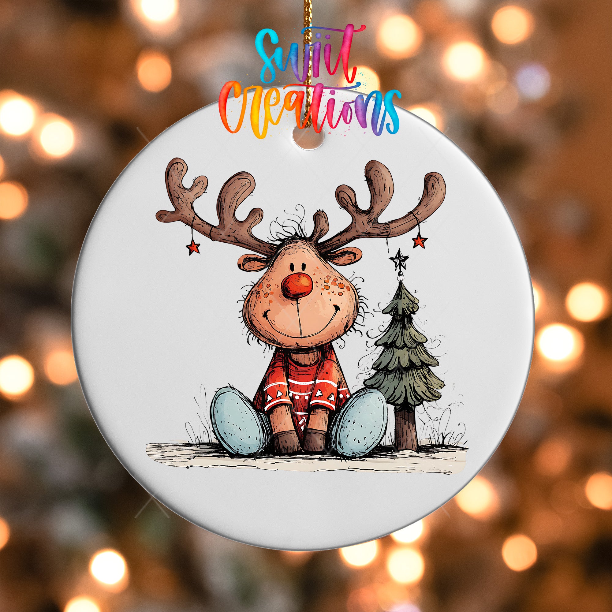 A white ornament with a cartoon reindeer wearing a red sweater and sitting next to a Christmas tree.