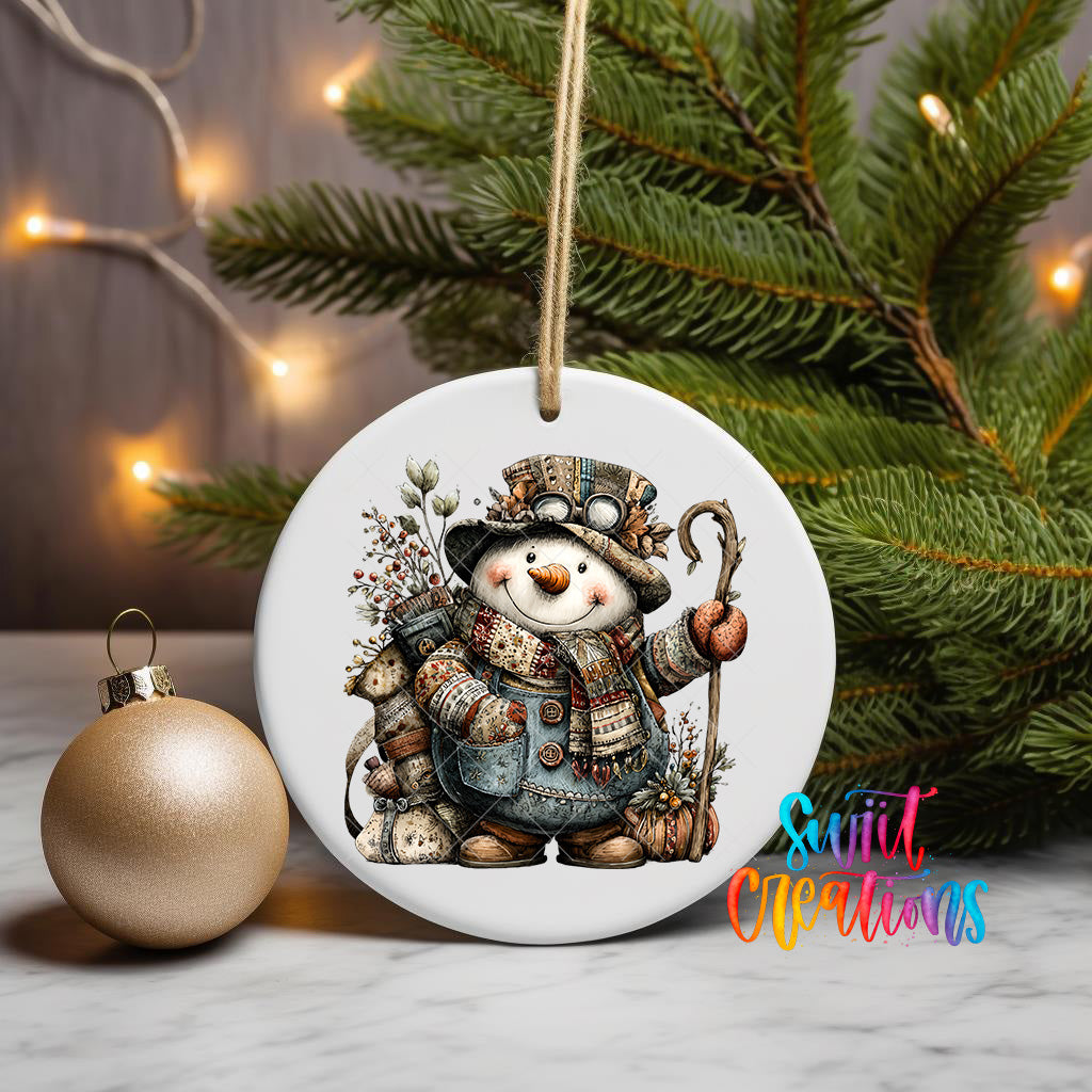 A white ceramic ornament featuring a snowman with a carrot nose and a staff, surrounded by pine branches and a gold ornament, with a string of lights in the background.