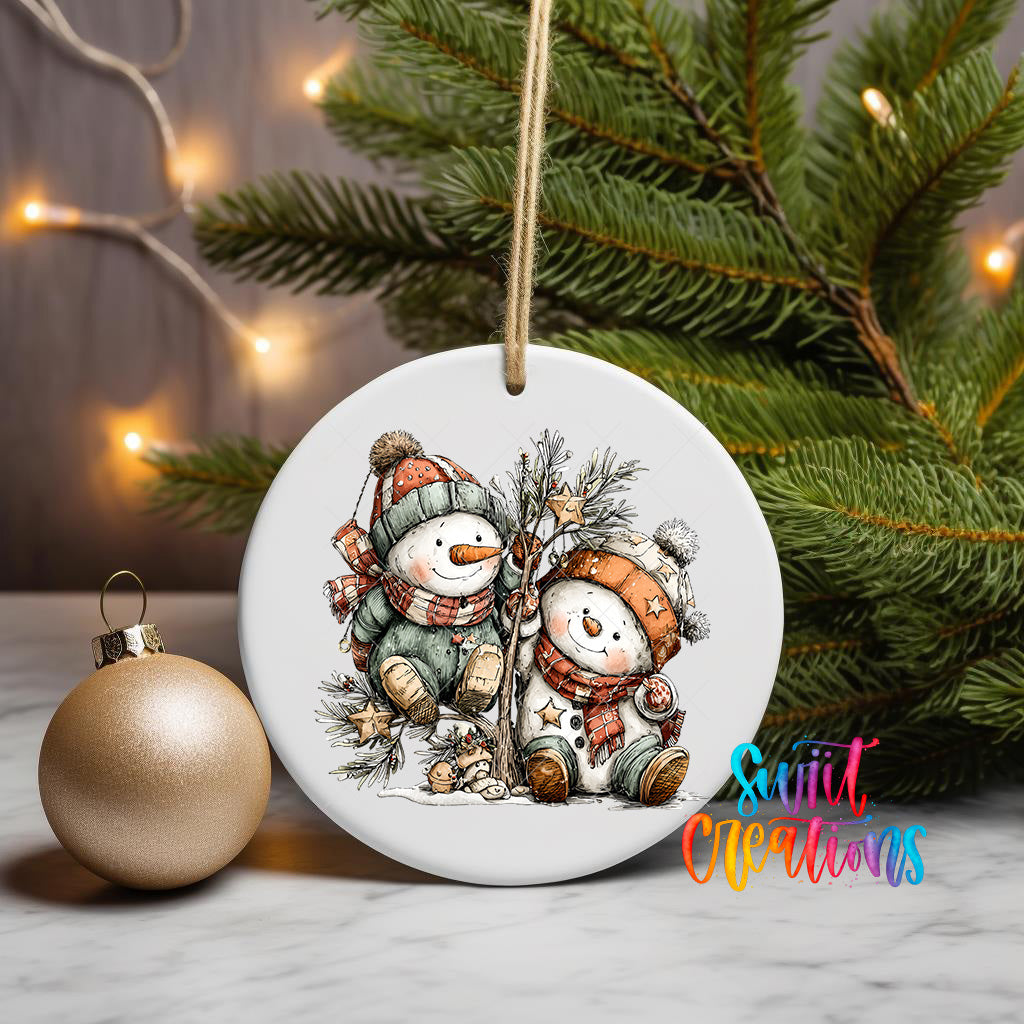 A white ceramic ornament featuring two snowmen sitting on a branch, surrounded by Christmas decorations and a gold ornament, with a Christmas tree in the background.