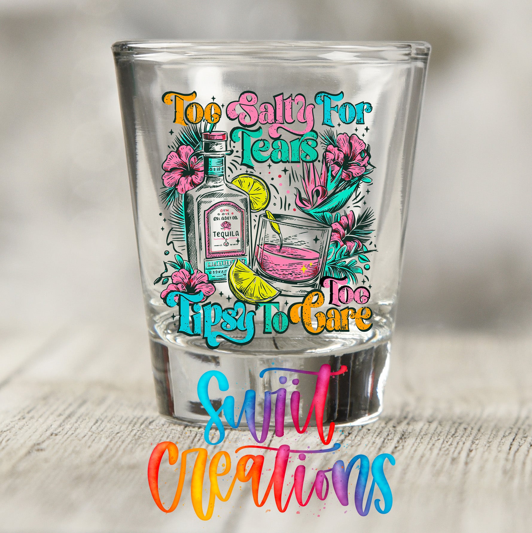 A colorful shot glass with a graphic design featuring a tequila bottle, a tequila glass, and tropical flowers, along with the text "Too salty for tears, tipsy to care" and "Sweet creations".