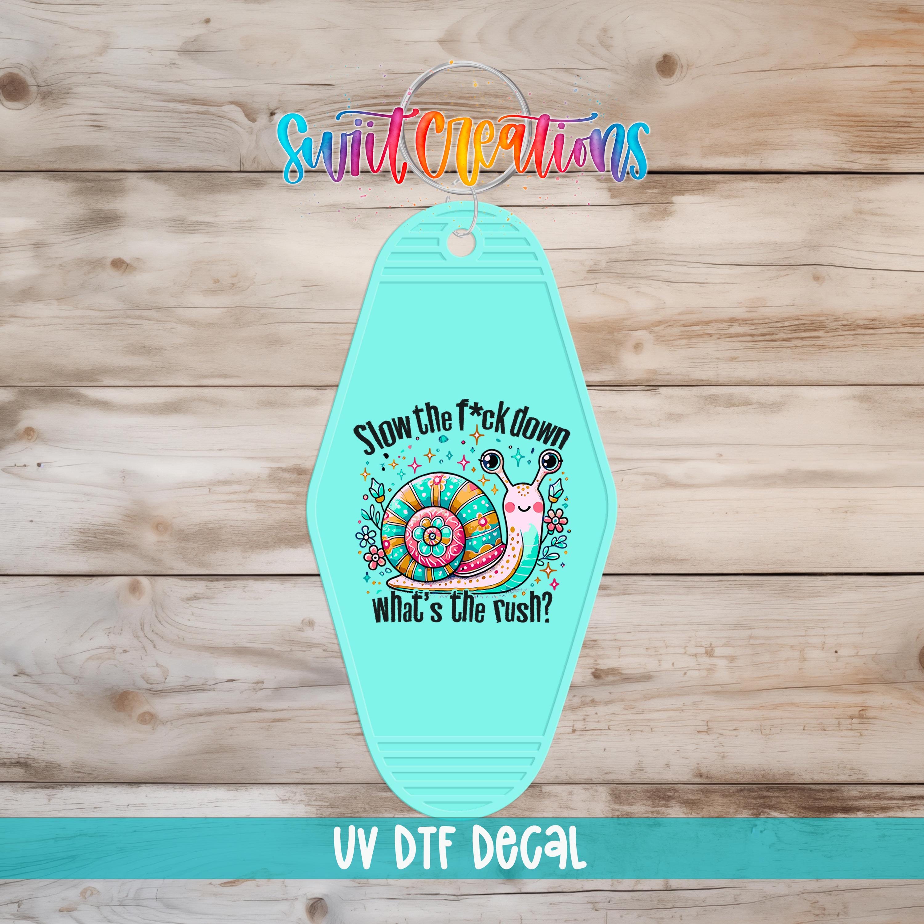 A turquoise keychain with a colorful snail design and text that says "Slow the fuck down what's the rush?"