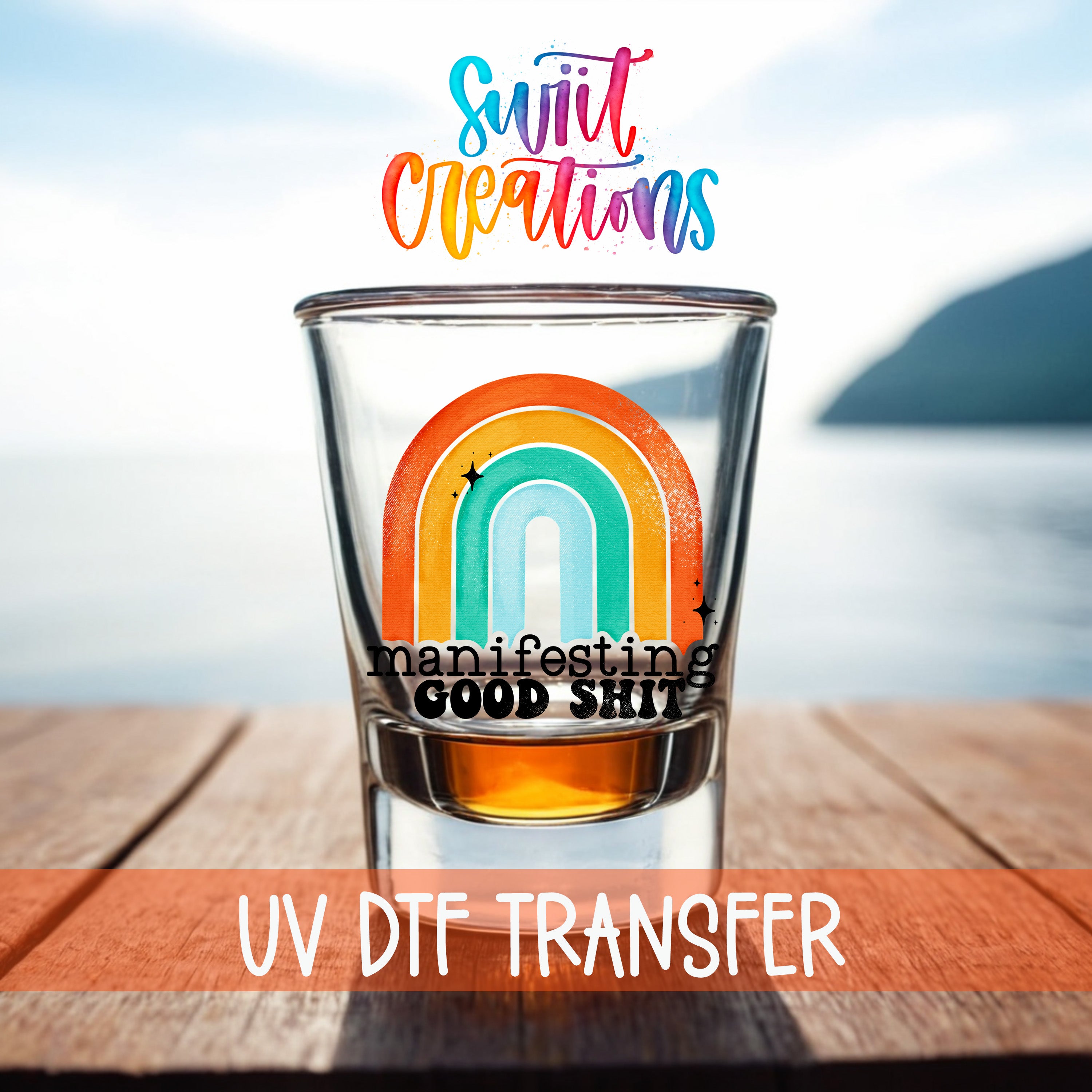 A shot glass with a rainbow graphic and the text "Manifesting GOOD SHIT" printed on it, placed on a wooden surface with a body of water in the background.