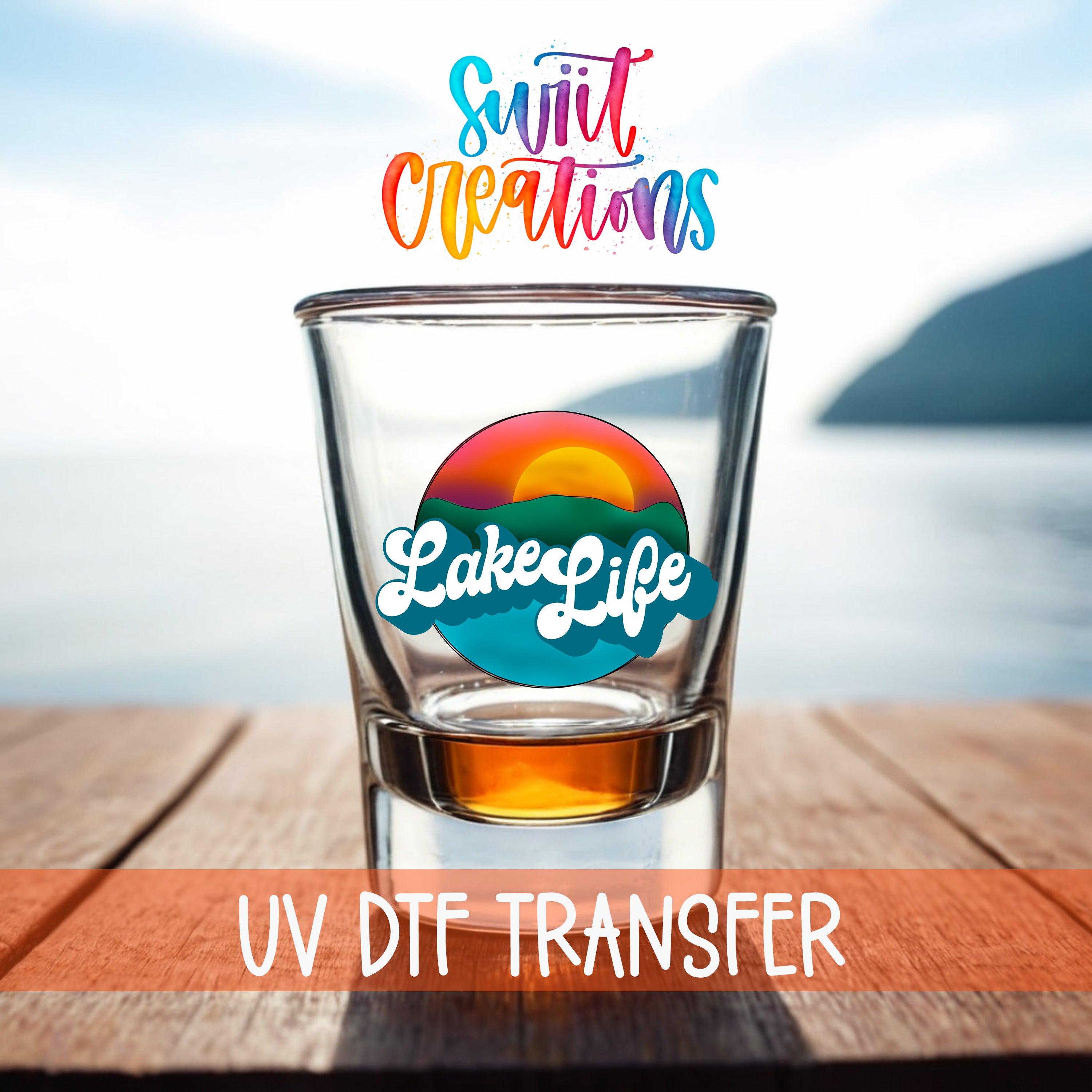 A shot glass with a colorful design on it, placed on a wooden surface with a scenic background of a body of water and a mountain.