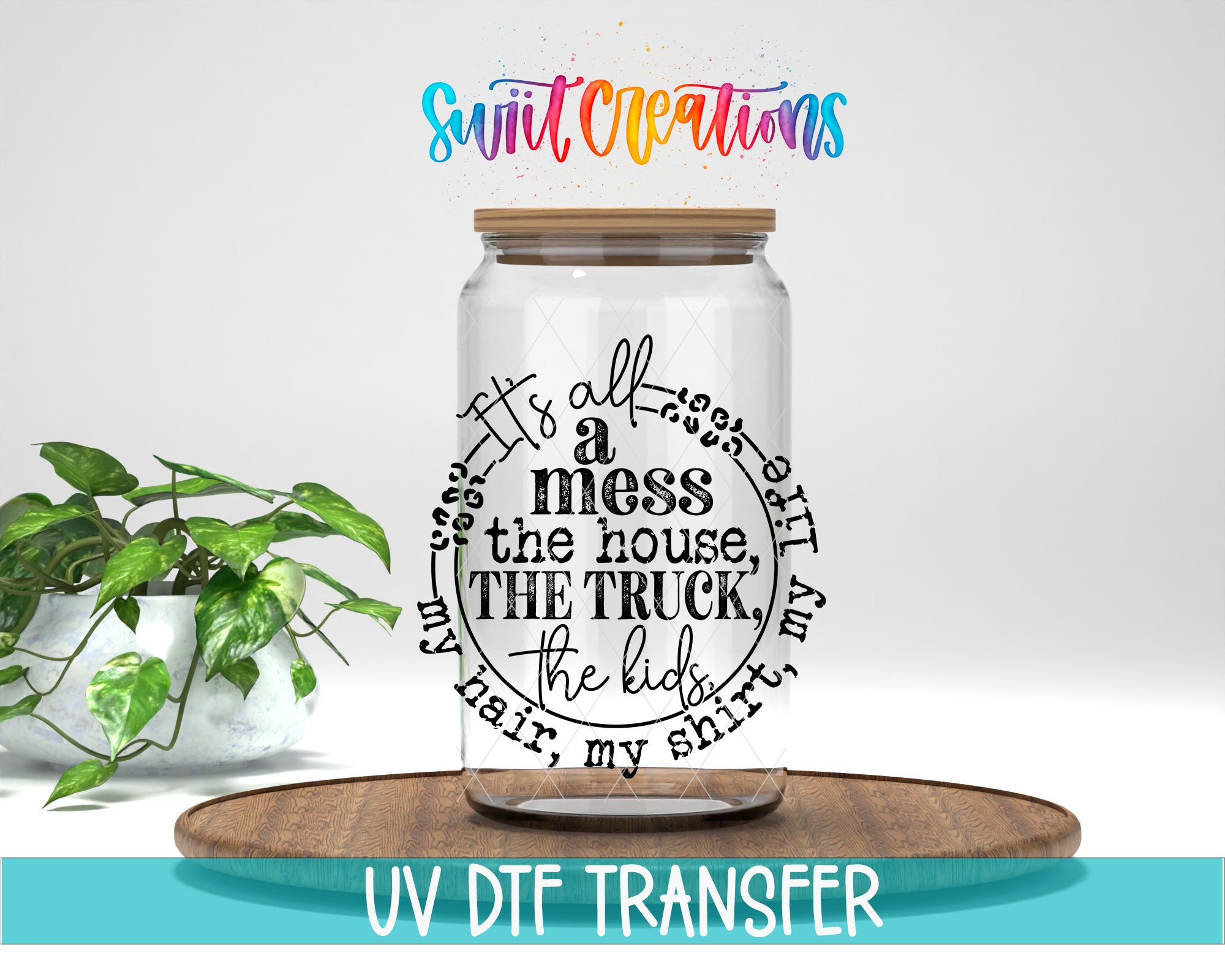 A clear glass jar with a wooden lid, featuring a hand-lettered quote that says "It's all a mess the house, the truck, the kids, my hair, my shirt, my life". The jar is placed on a wooden tray, and there is a potted plant in the background.