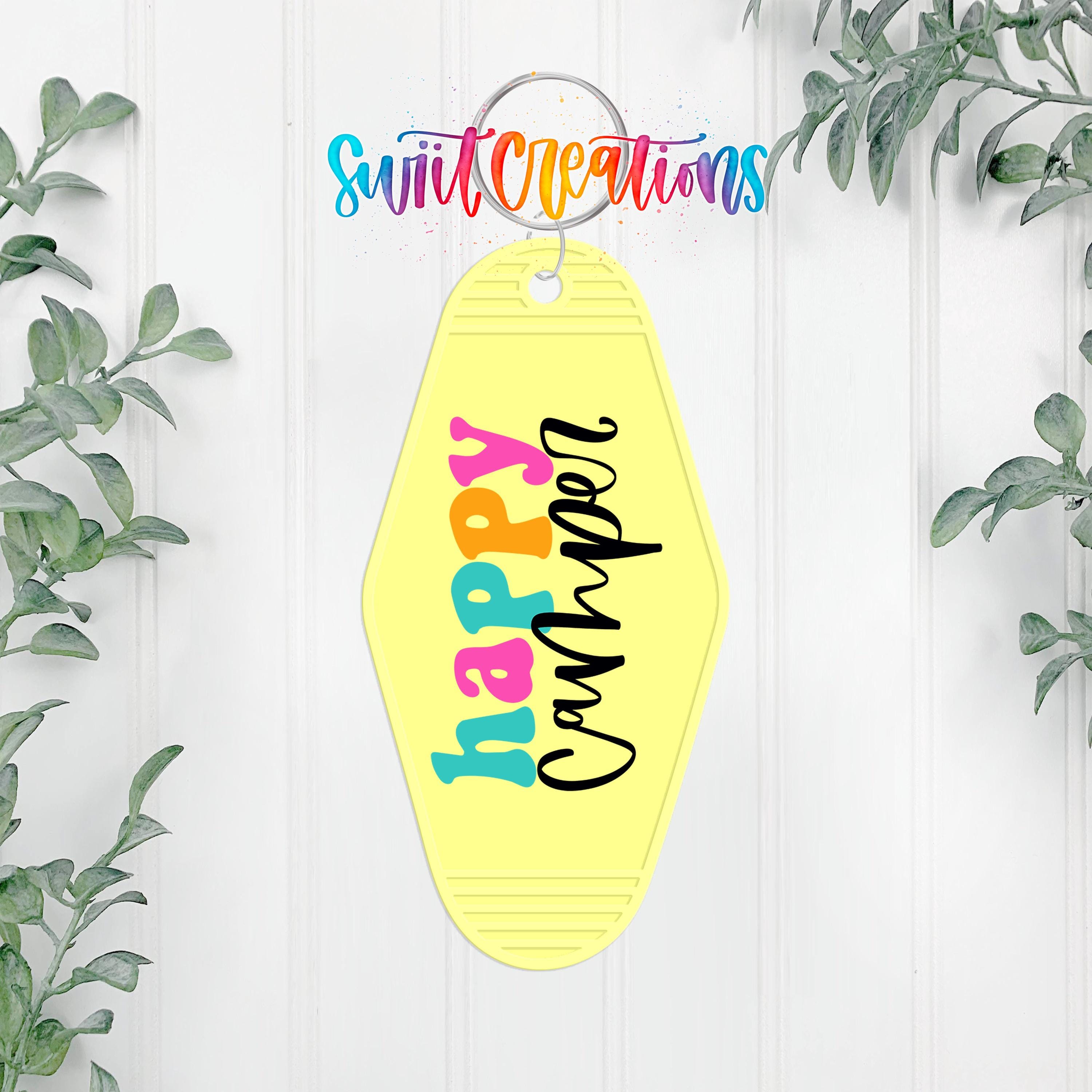 A yellow keychain with the text "happily camped" printed on it, hanging on a white wall with greenery in the background.