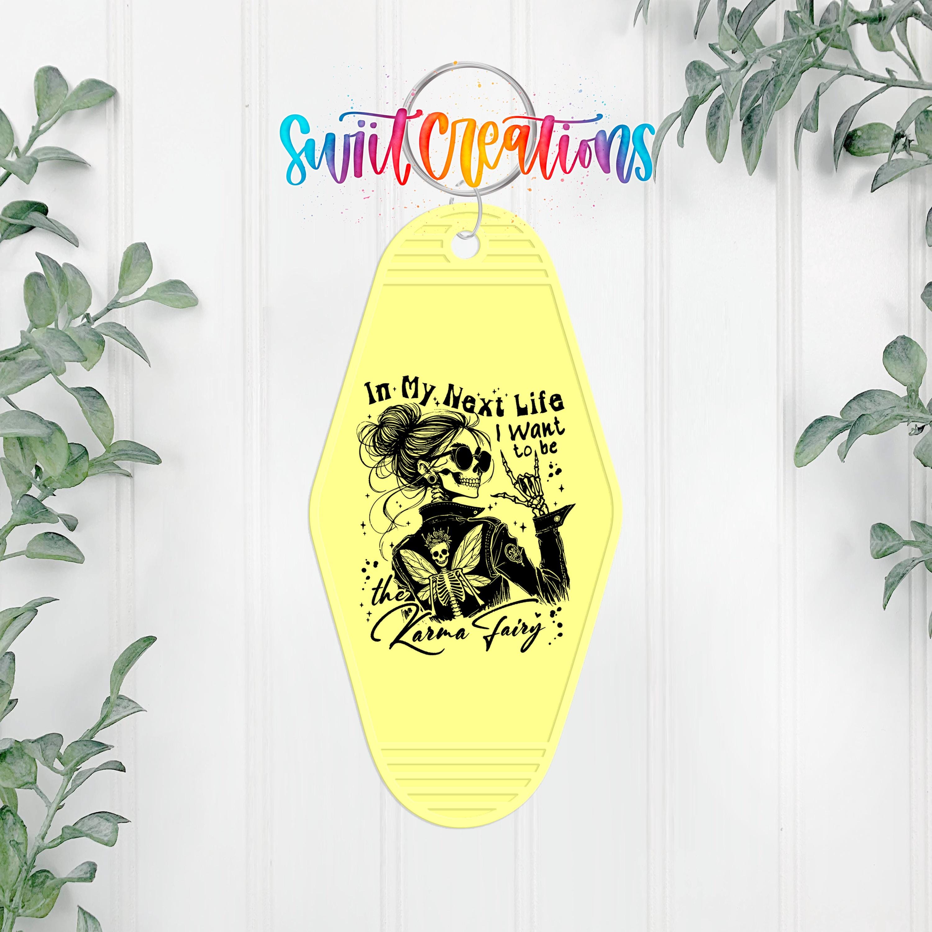 A yellow keychain with a black and white illustration of a woman with a bird on her shoulder, along with the text "In My Next Life I Want to Be the Karma Fairy".
