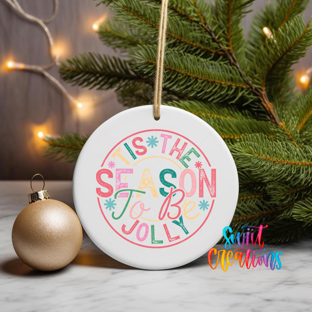 A white ceramic ornament with a colorful design hangs on a Christmas tree, surrounded by greenery and twinkling lights.