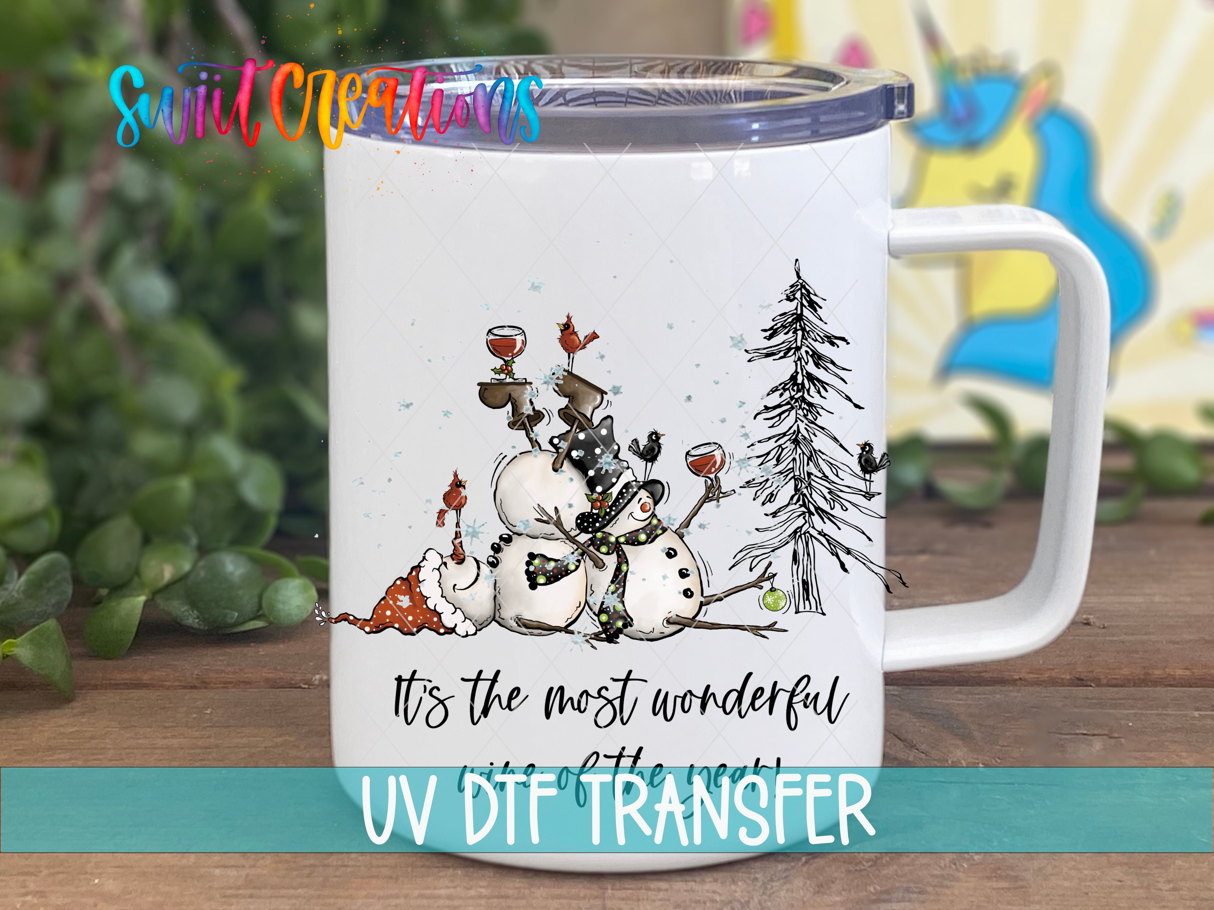 A white mug with a winter scene design featuring a snowman, a pine tree, and birds. The mug has a handle and a lid, and there is text that says "It's the most wonderful UV Df Transfer".